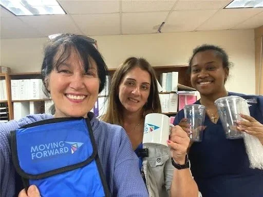 Three women smiling and holding clear plastic cups in an indoor setting, with bookshelves in the background.