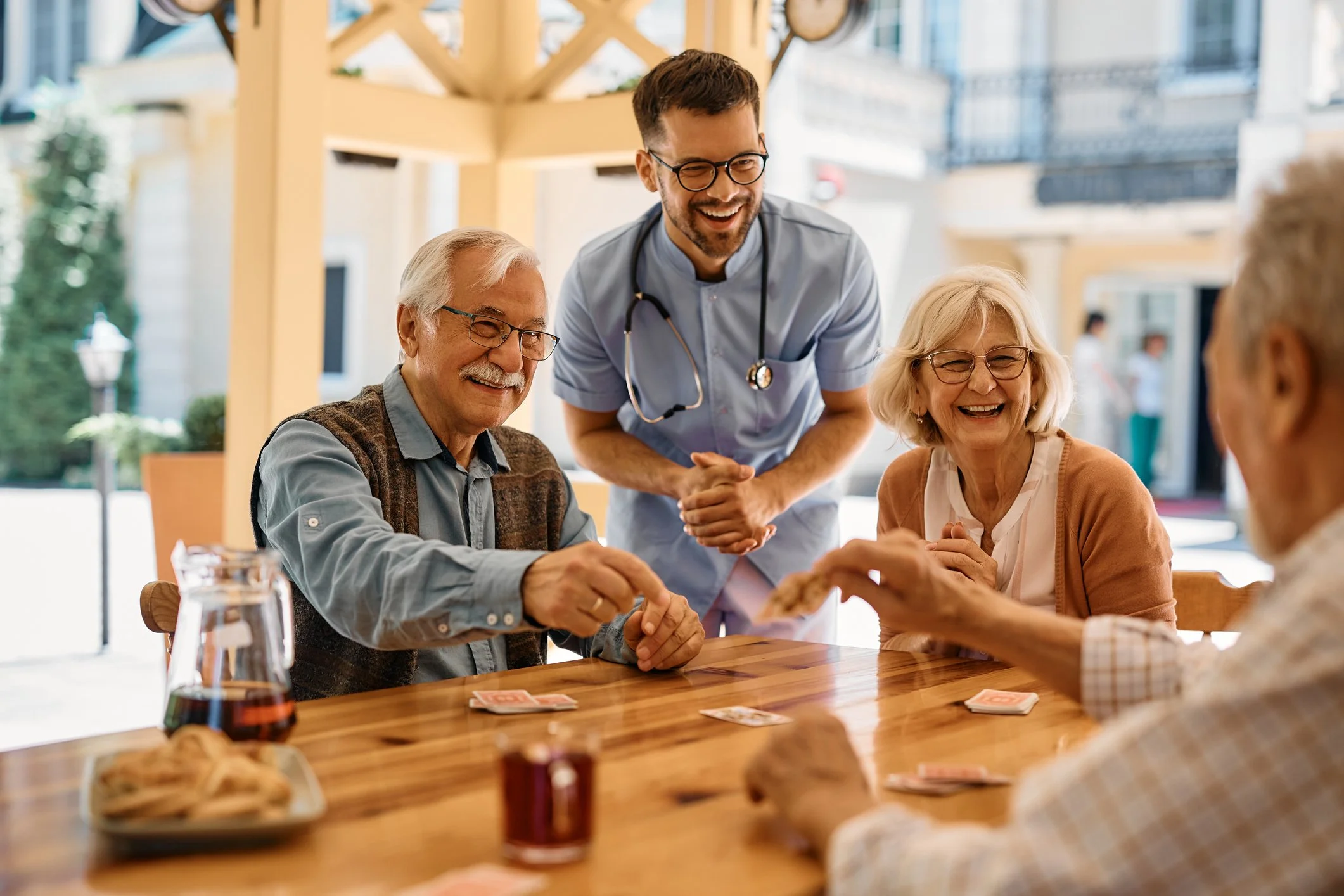 A group of elderly friends and a young male nurse playing cards at a wooden table in an outdoor patio, smiling and laughing while the nurse serves tea or coffee. There is a plate of cookies and a pitcher of tea on the table.