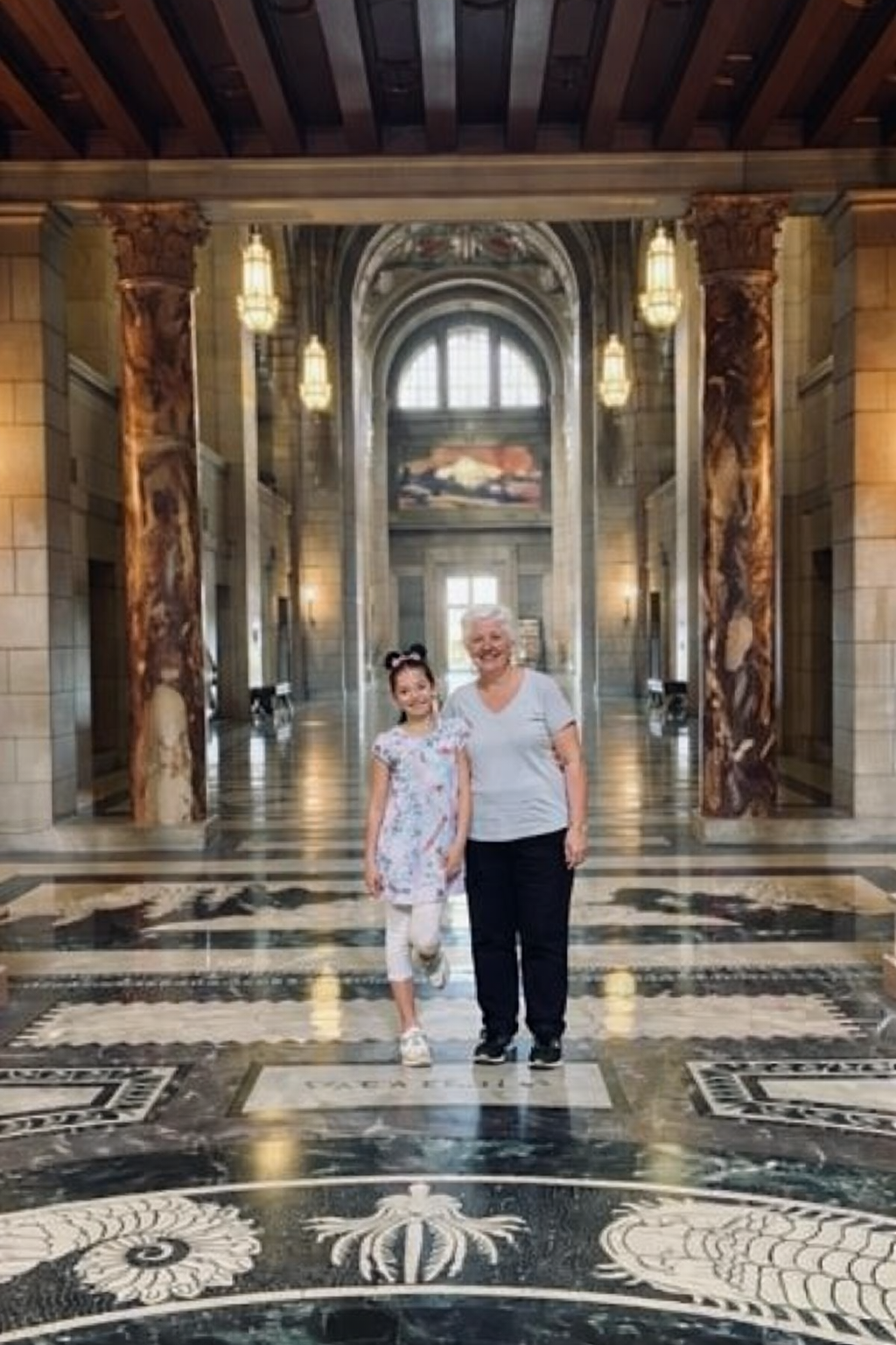 A young girl and an older woman standing together inside a grand, ornate building with marble floors, columns, and large windows.
