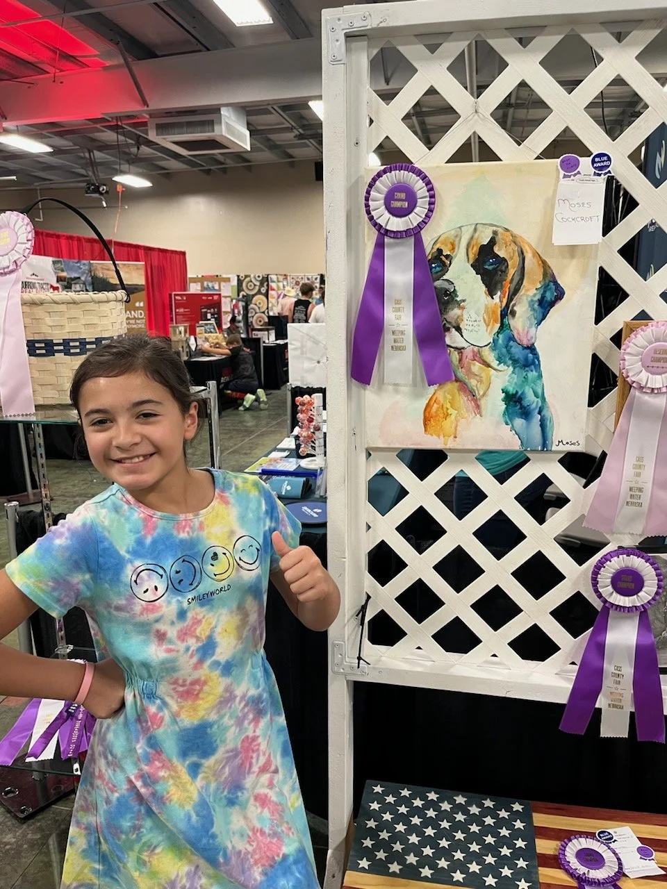 A young girl smiling and giving a thumbs up at an art and craft fair, standing next to a watercolor painting of a dog, which has award ribbons displayed nearby.