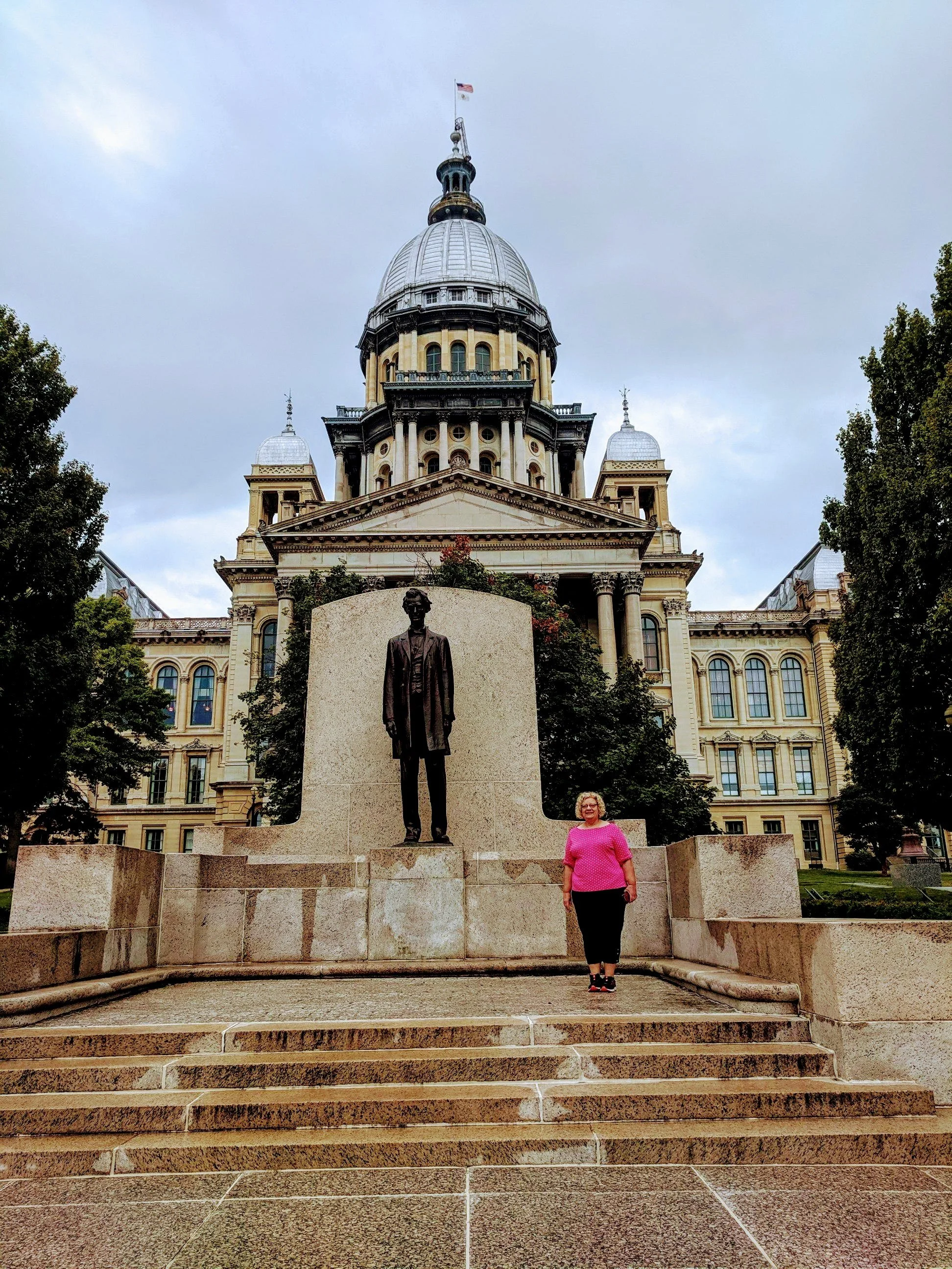 A woman in a pink shirt standing in front of a statue of a man in a suit outside a large historical government building with a dome, surrounded by trees.
