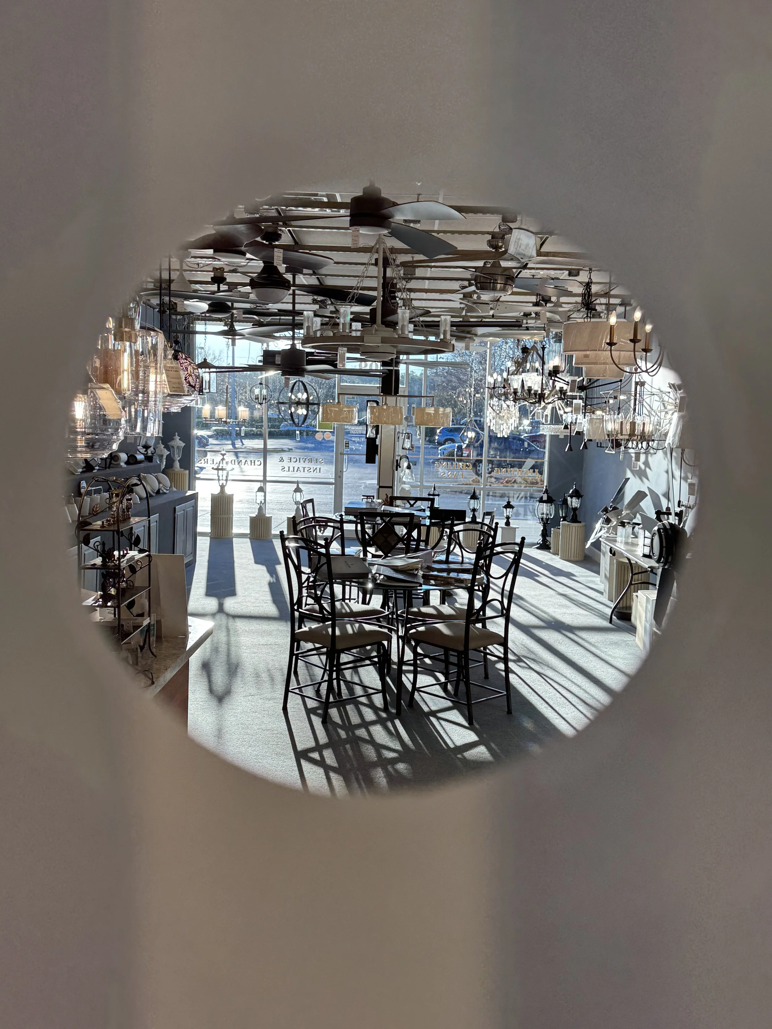 Interior of a furniture store, viewed through a circular opening, with tables, chairs, chandeliers, ceiling fans, and lamps, and large windows at the front letting in natural light.