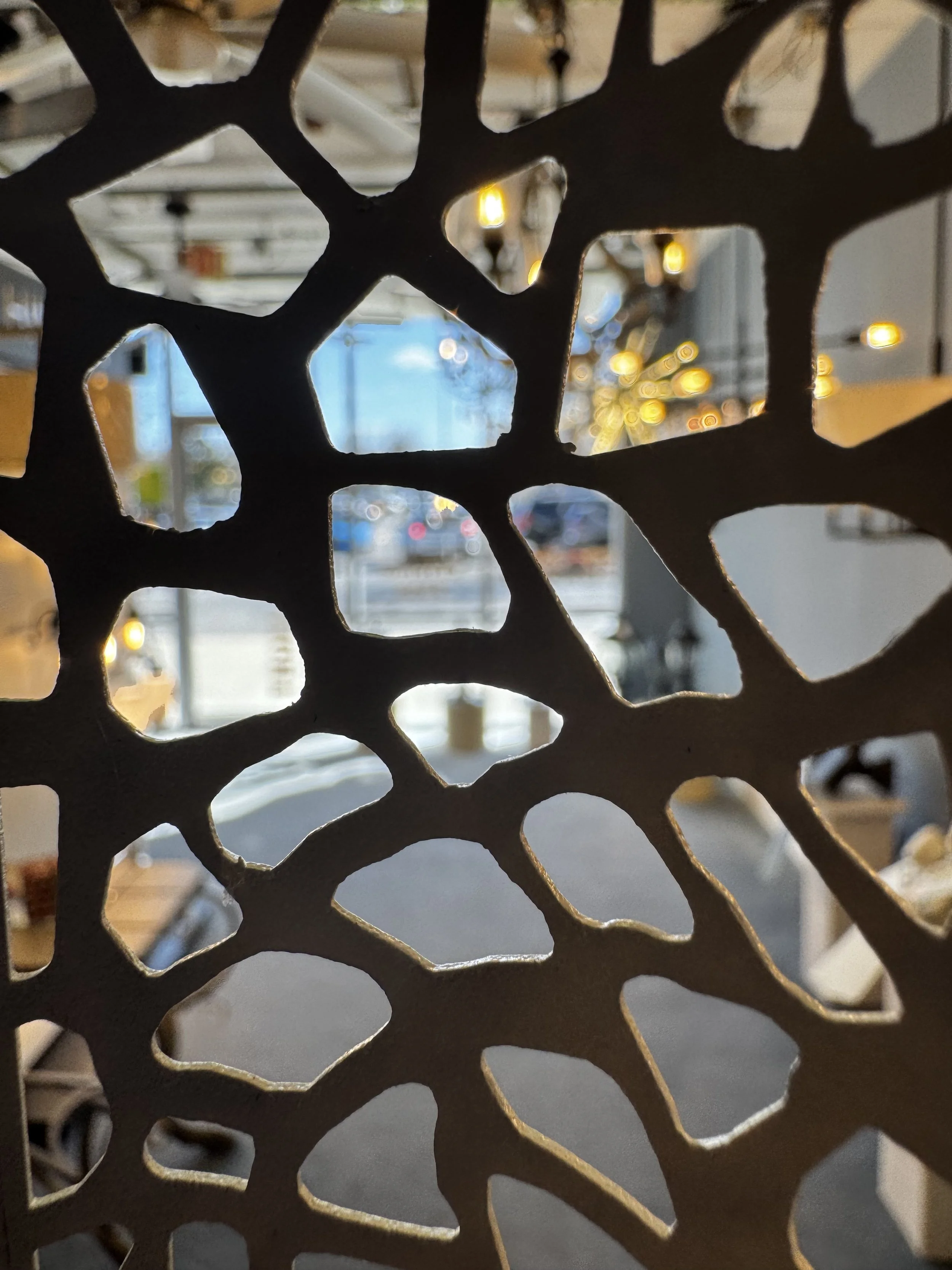 Close-up view through a decorative, honeycomb-patterned metal screen with warm yellow hanging lights visible in the blurred background outside.