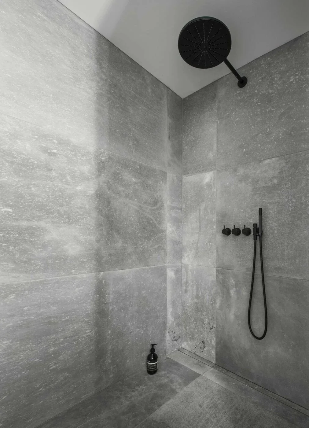 Mustavalkoinen moderni suihkuhuone, jossa harmaa kivireunainen seinä ja musta suihkusetti, including rain showerhead and hand-held shower, sekä pieni musta saippuapullo lattialla.