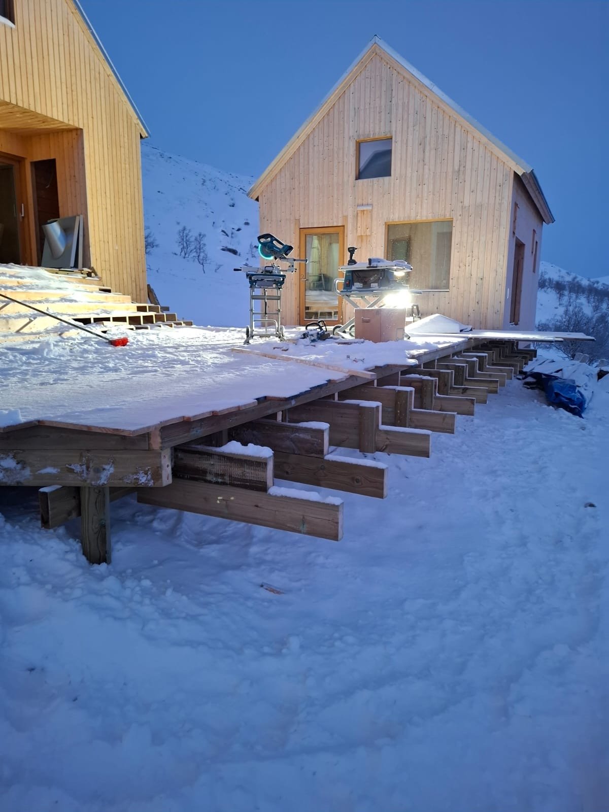Luminen, puinen talo rakennusvaiheessa lumisessa ympäristössä, rakentamistyökalut ja työkoneet kuvassa.