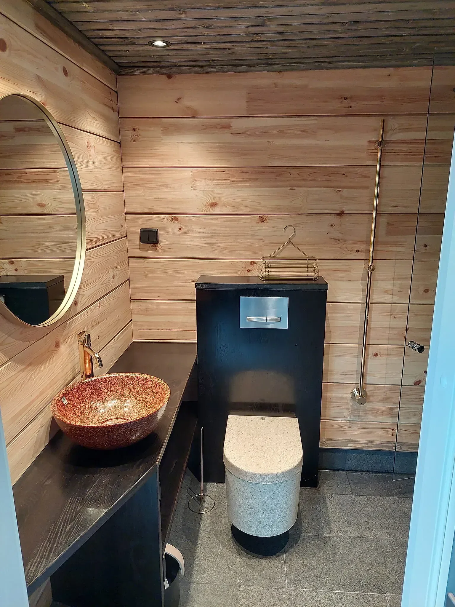 Kuva uudesta kylpyhuoneesta, jossa puinen seinä ja katto. Vasemmalla on pesuallas ja peili, keskellä on musta ovi ja pieni kylpyamme. Oikealla on suihku ja lipastokaappi.