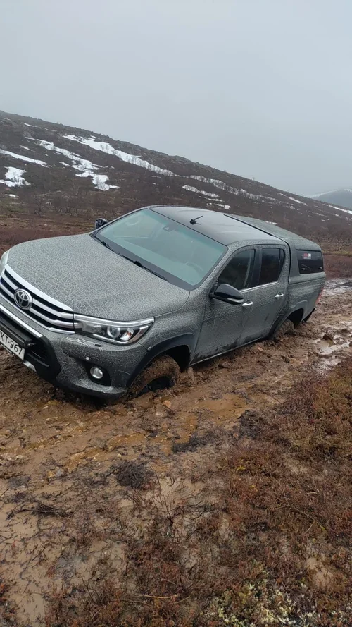 Musta pickup-auto ontuu syvälle mutaan ja on osin peittynyt veteen, maasto on epätasainen ja mutainen, ja tausta on vuoristomainen. Sade tai sumu näkyy taustalla. Rakennusliike Utsjoki (Construction company Utsjoki)Rakennuspalvelu Inari 