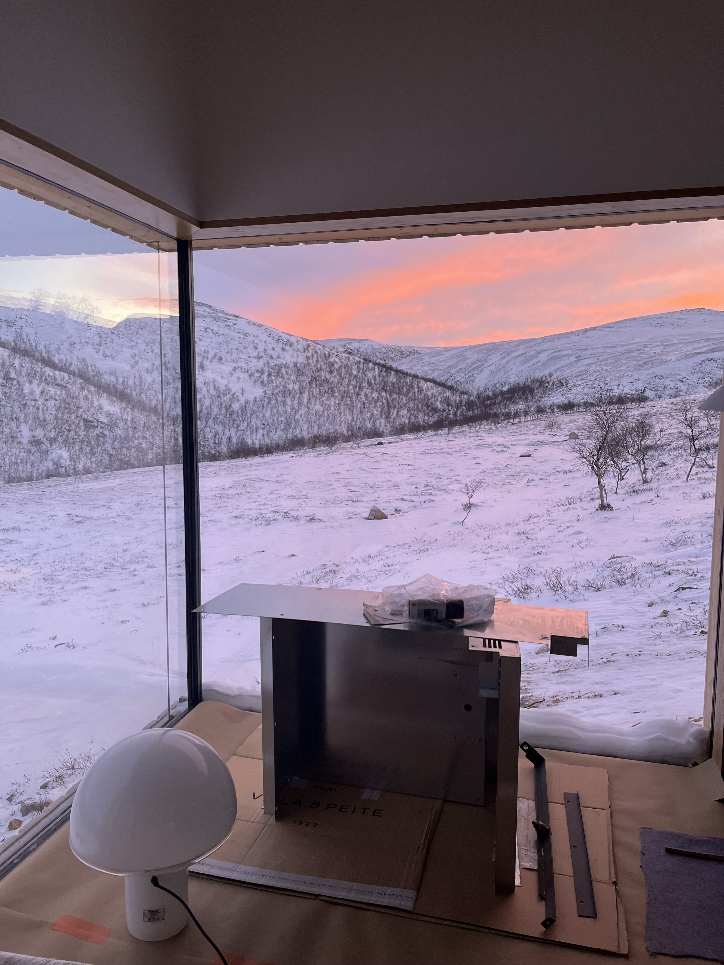 Talo ikkunasta avautuva luminen vuoristomaisema auringonlaskun aikaan, sisällä pöytätaso ja osia rakennusprojektista.