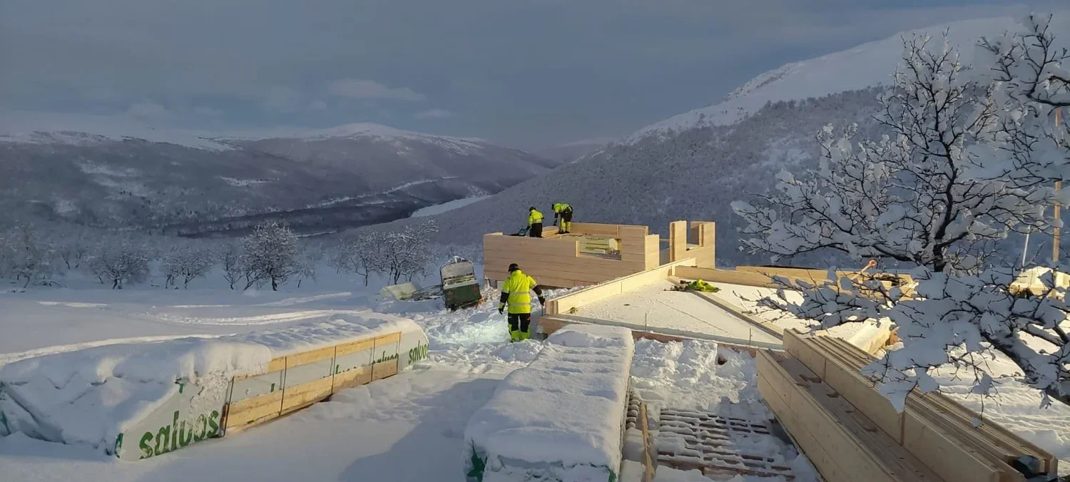 Rakennustyömaa lumisessa maisemassa, työtekijöitä rakentamassa puutaloa talven keskellä, taustalla mäkisiä vuoria ja lumisia puita. Rakennusliike Utsjoki (Construction company Utsjoki)
Rakennuspalvelu Inari (Construction service Inari) Urakoitsija 