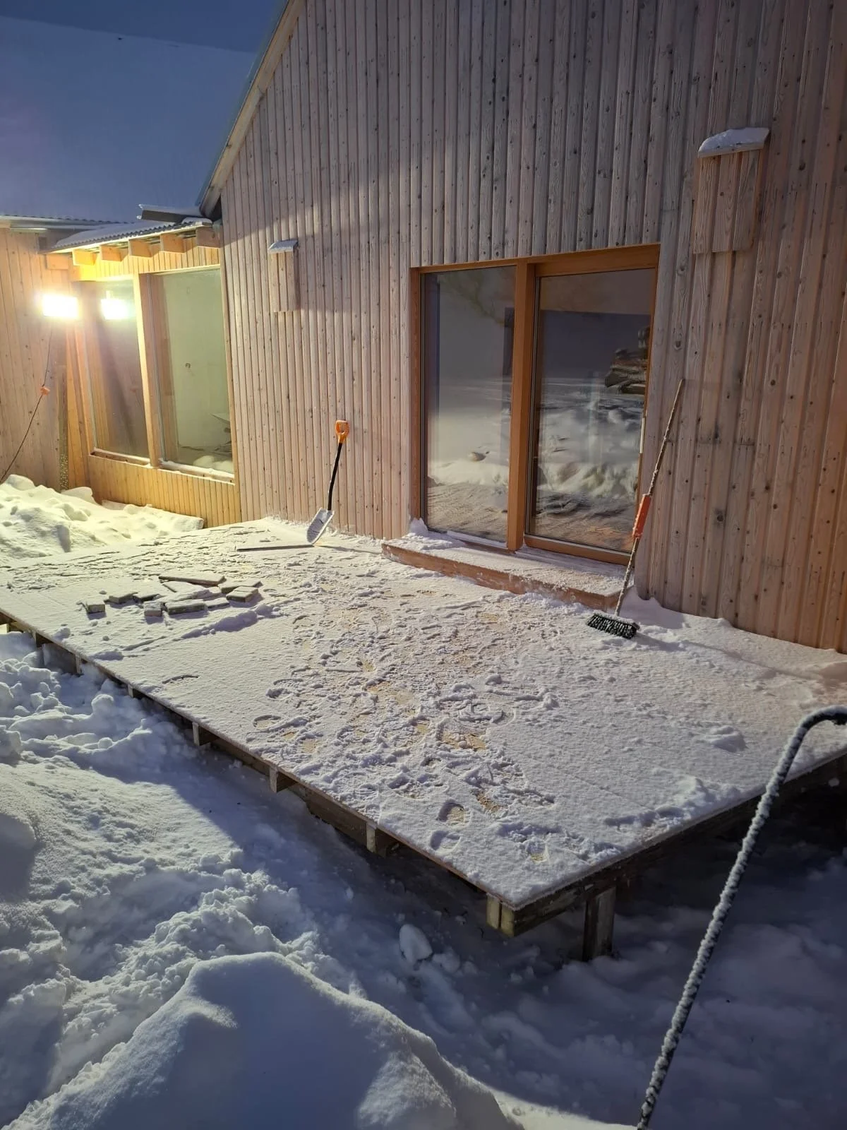 Rakennus, jonka seinät on päällystetty puulla, ja lasiovet näkyvät ulkona lumisessa ympäristössä. Työvälineet kuten lumiharja ja lapiot ovat näkyvillä, ja rakentamisen jäljet ovat pinnalla.