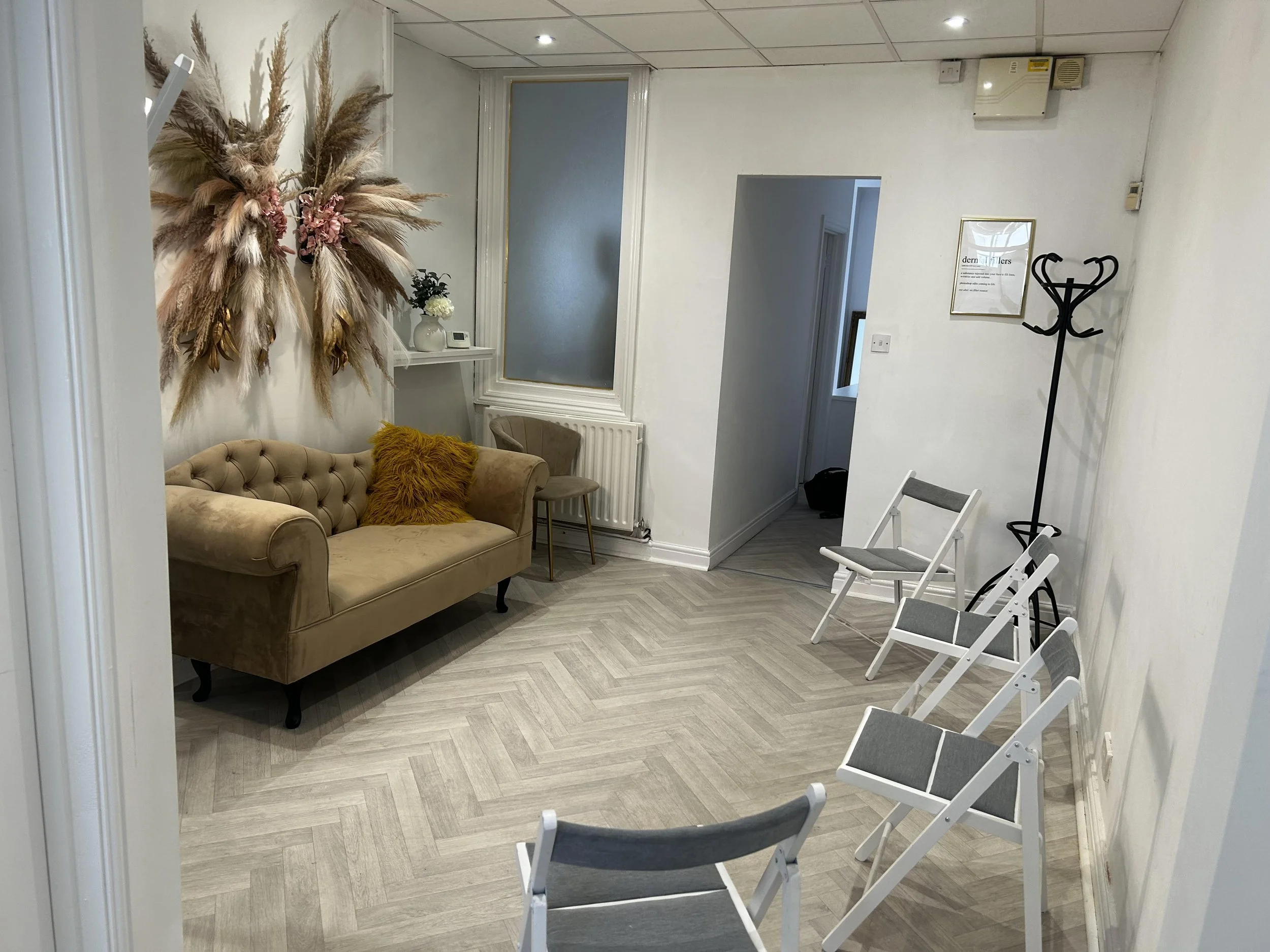 An interior room with a beige tufted sofa with a mustard-colored pillow, a small beige chair, a wall decorated with a large dried flower arrangement, a framed sign, and a coat rack. White folding chairs are arranged in a row.