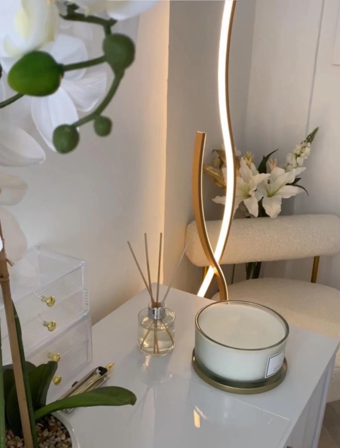 Decorative vanity table with reed diffuser, candle, orchid flowers, and a modern illuminated mirror in a bedroom.