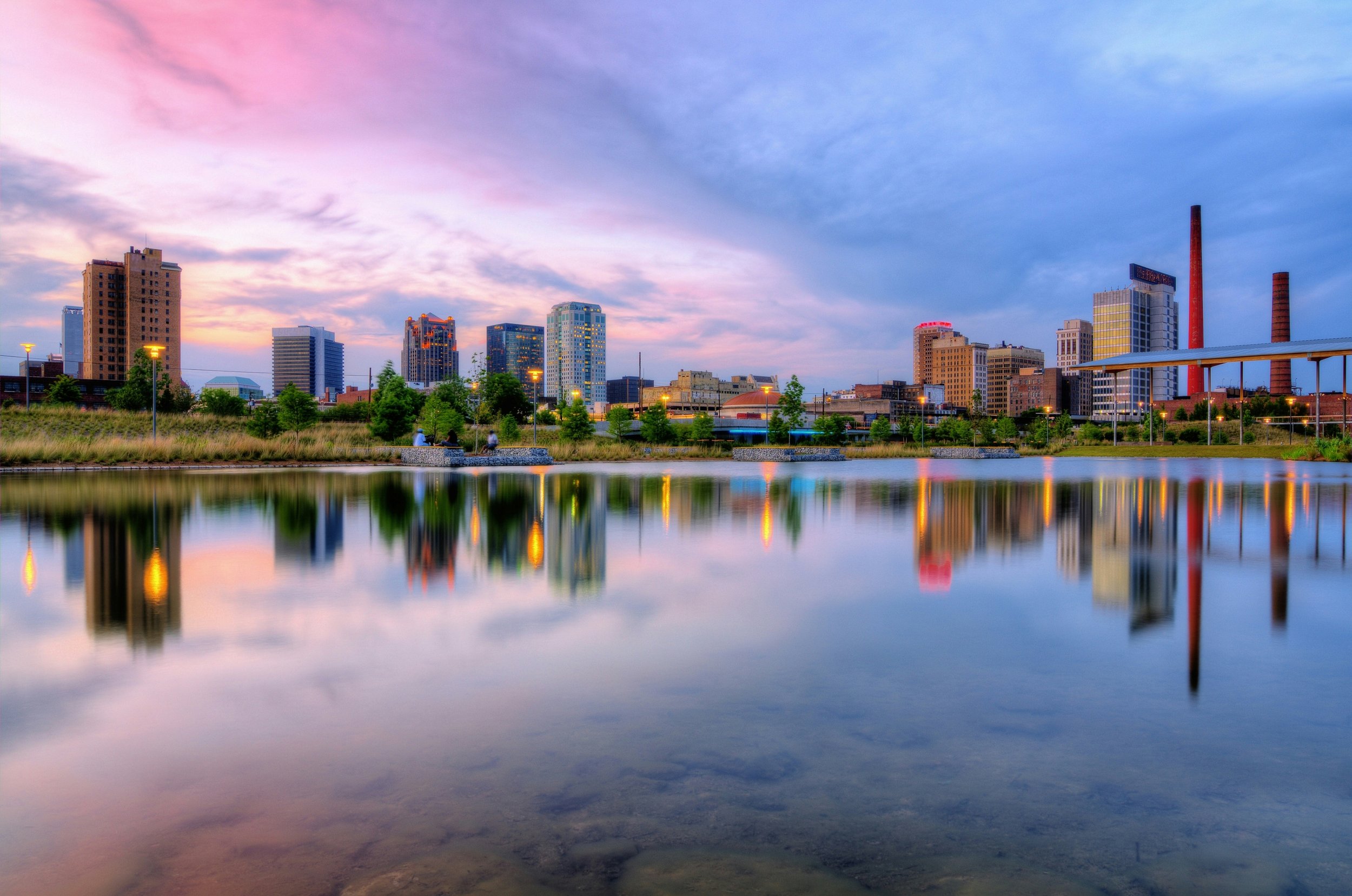 City skyline with tall buildings and a river or lake in the foreground at sunset or sunrise, with colorful sky and reflections on the water.
