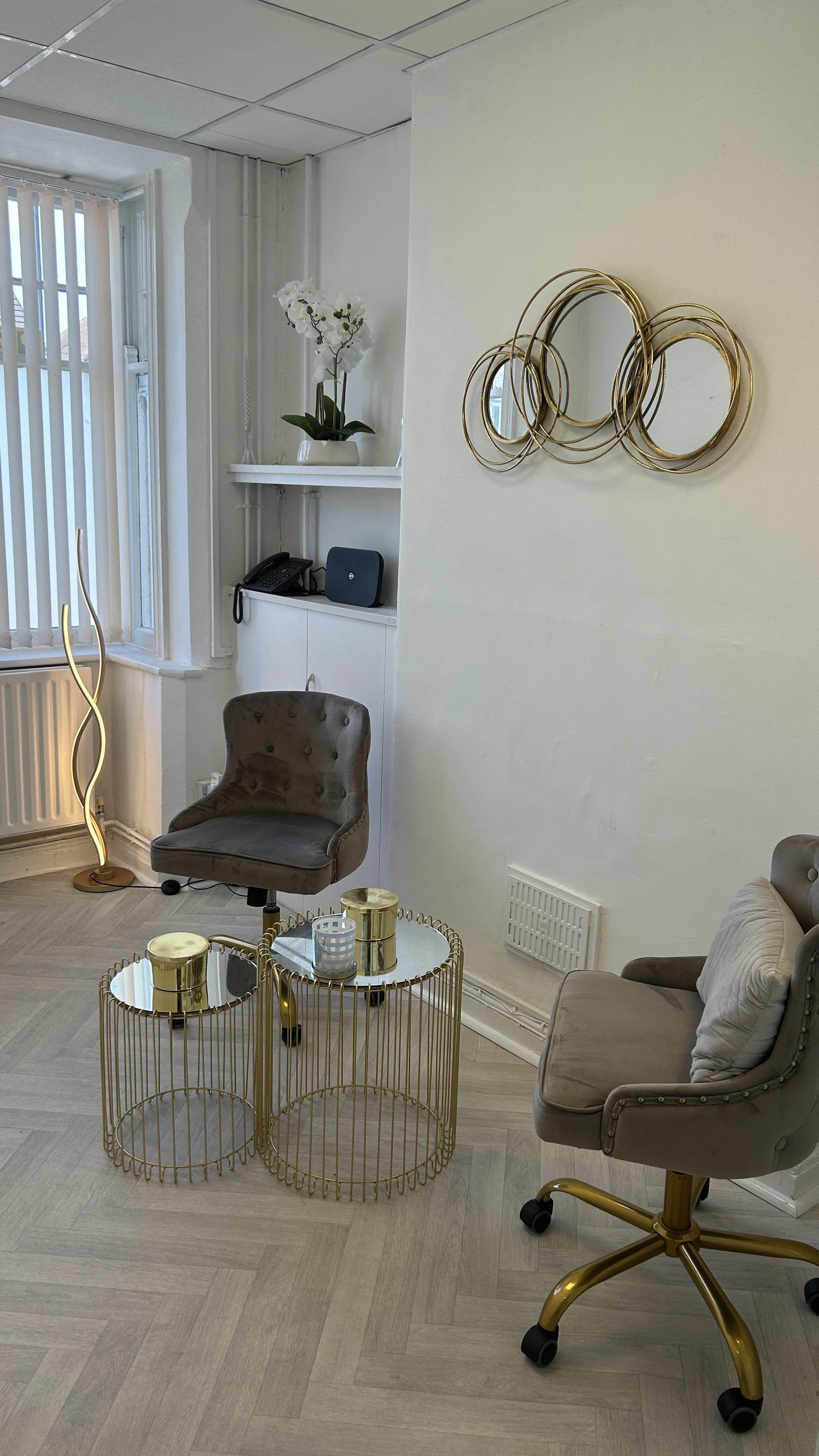 Living room with beige and gold accents, including two beige armchairs with gold legs, a gold wireframe coffee table with metallic containers, a black chair, and decorative mirrors and a white orchid on a shelf, near a window with vertical blinds.
