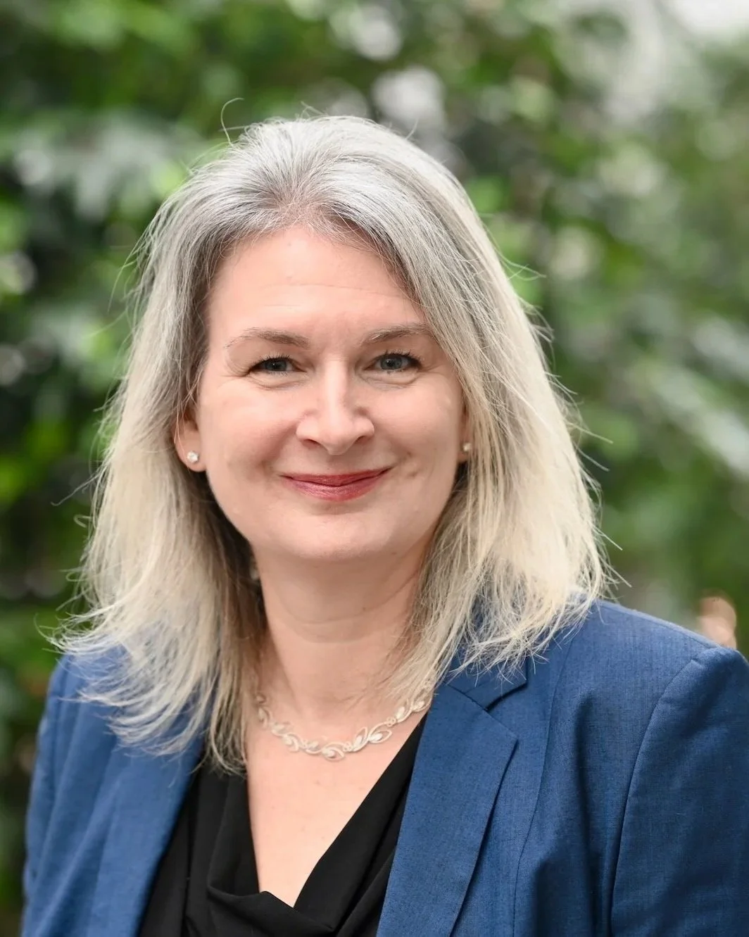 A photo of Kristin Wildman Shirahama, a founding partner at Fides Law. She has long gray hair, is smiling, and wears a blue blazer and black top, with a necklace and earrings, standing outdoors against a blurred green background.