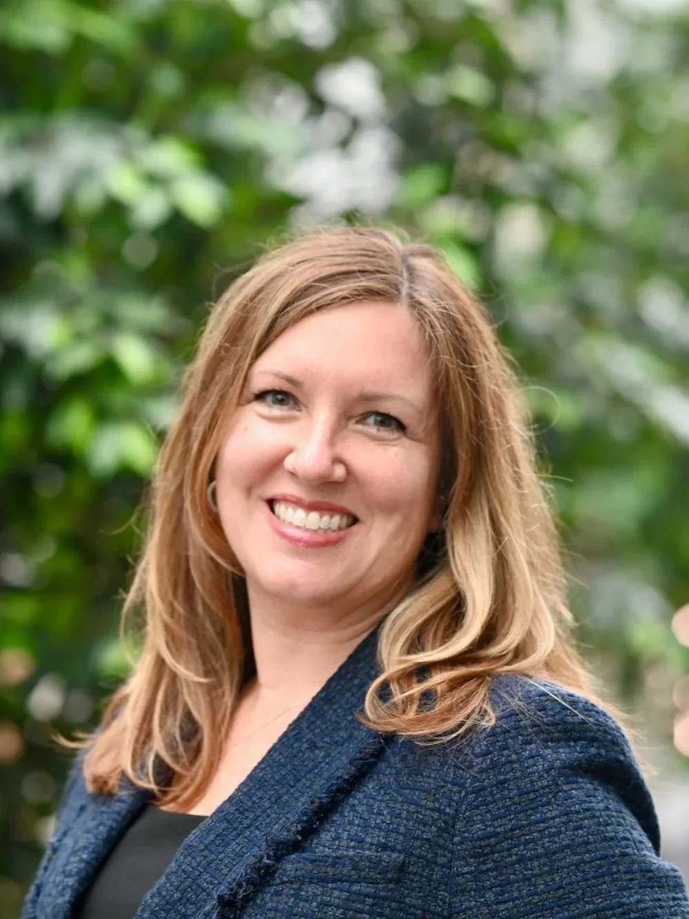 A photo of Rebecca MacGregor, a founding partner at Fides Law. She has shoulder-length light brown hair, is wearing a dark blazer, and is standing outdoors against a background of green foliage.