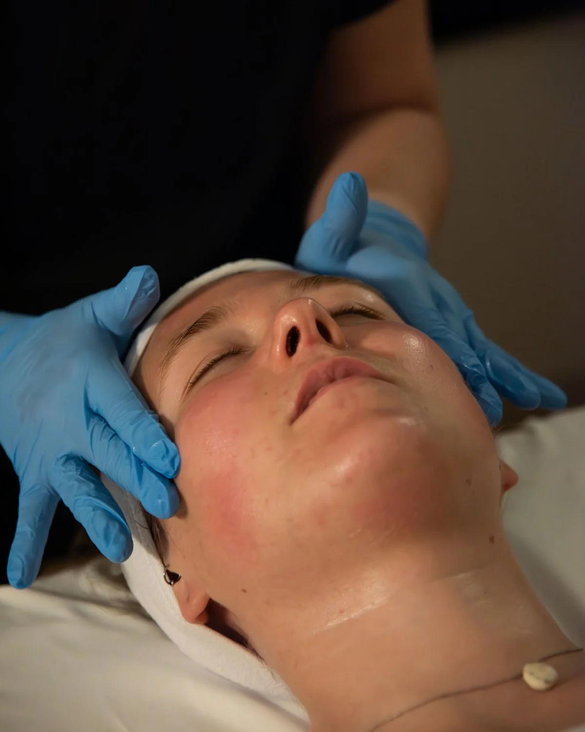 A person lying down with eyes closed receiving facial treatment from a professional wearing blue gloves.