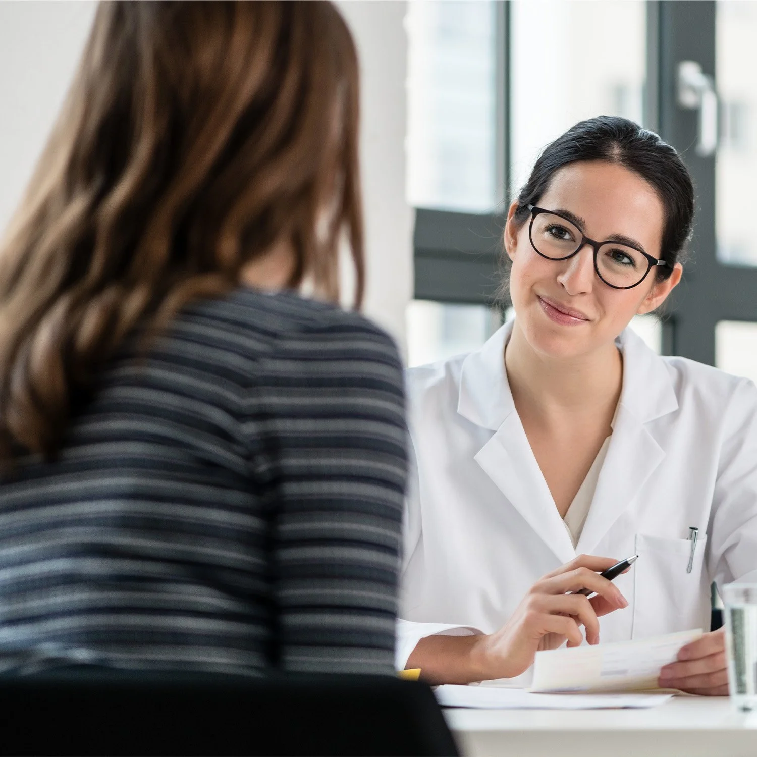 A female doctor with glasses in a white coat smiling and talking to a woman with brown hair and a striped shirt during an appointment in a bright medical office.