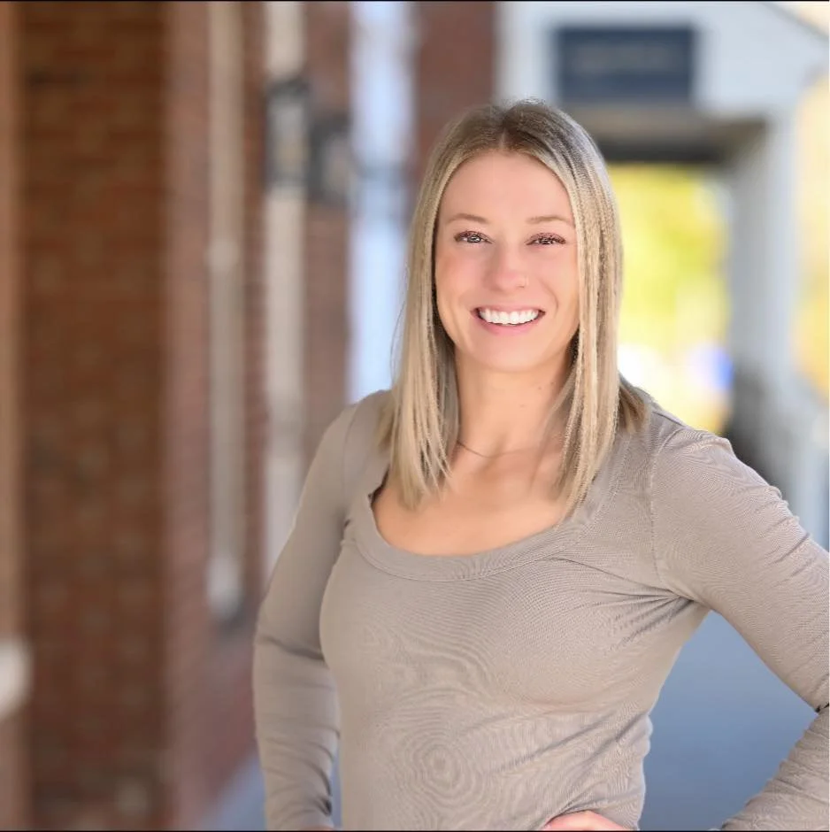 A woman with blonde hair smiling and standing outdoors in front of a blurred background of buildings and trees.