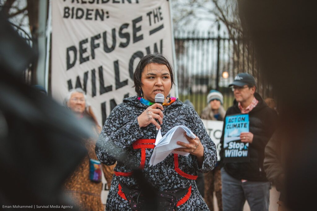 A woman speaking into a microphone during a protest, holding papers in her hand, with a large banner behind her that says 'Refuse the Will of the...' and other people holding signs in the background.