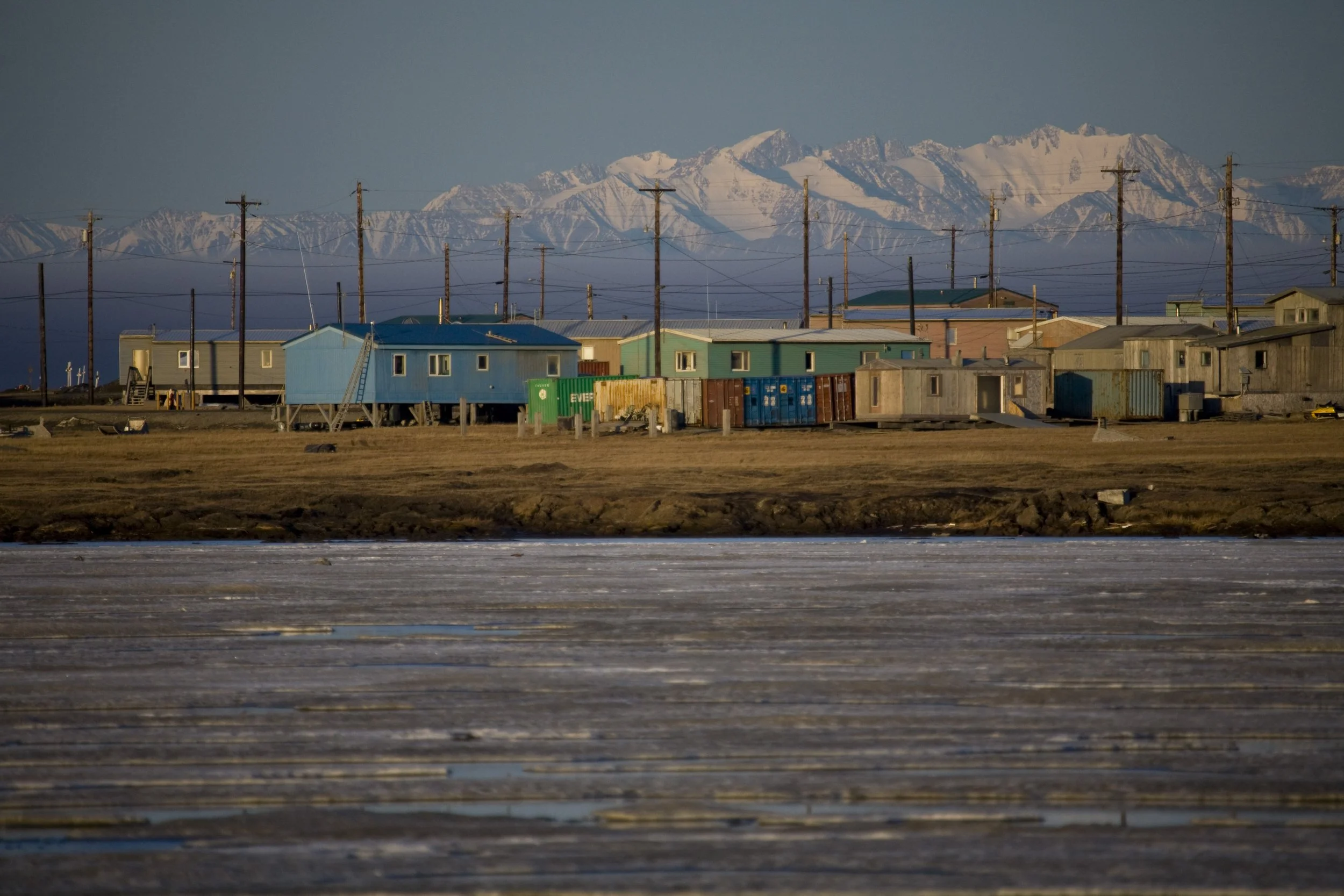 A row of colorful houses with utility poles, snow-capped mountains in the background, and a frozen water body in the foreground.