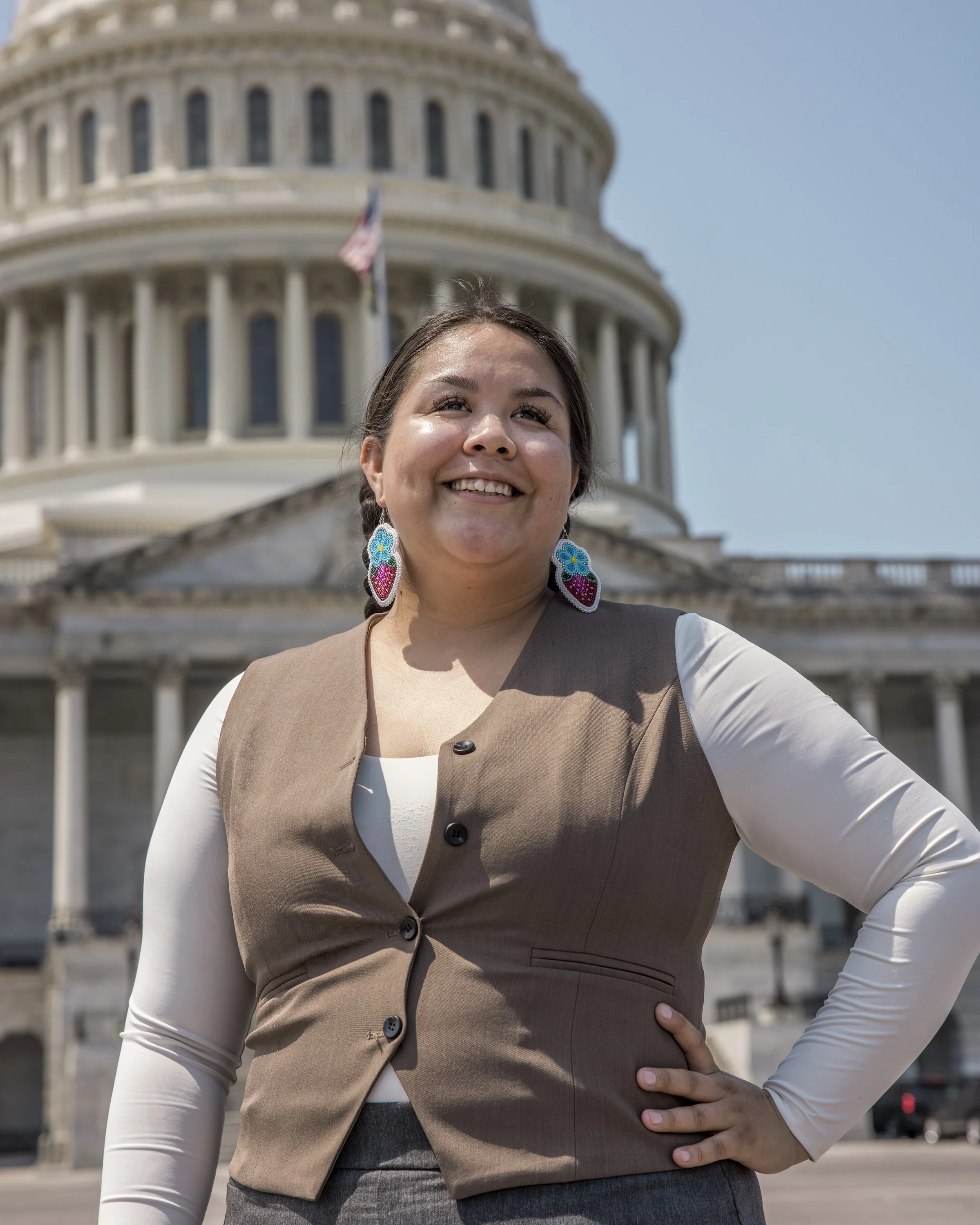 A woman standing in front of the U.S. Capitol building, smiling and posing with her hand on her hip, wearing colorful strawberry earrings and a beige vest over a white long-sleeve shirt.