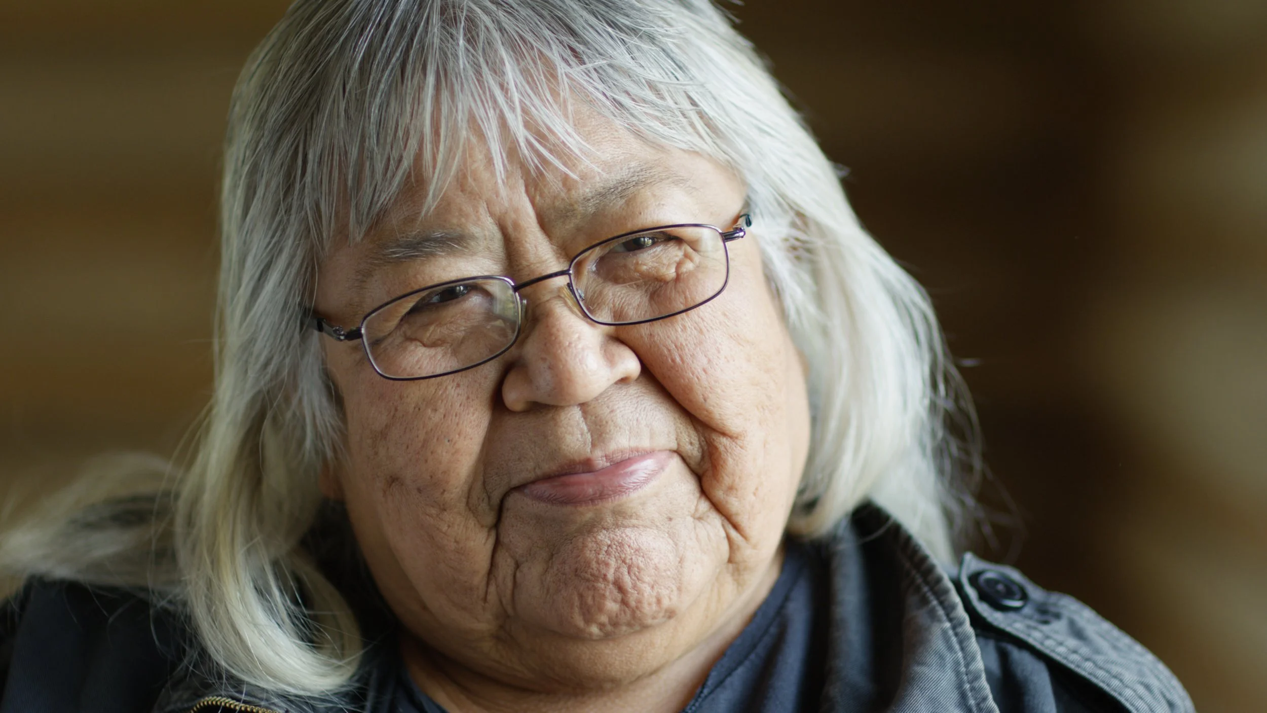 Close-up of an elderly woman with long gray hair, wearing glasses and a dark jacket, looking thoughtfully to the side.
