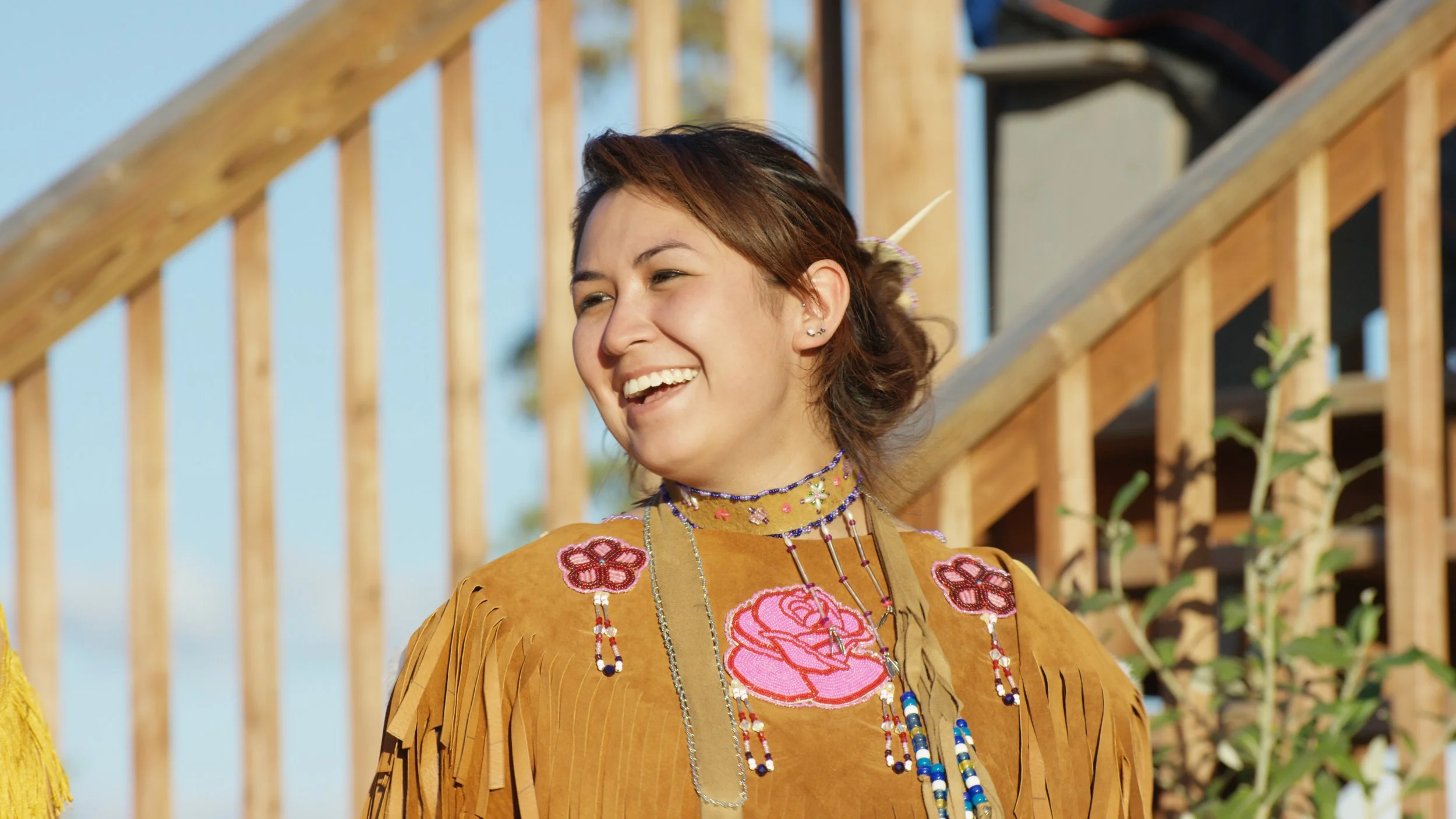 A young woman with brown hair in a loose bun, smiling outdoors, wearing a brown fringed suede top with floral embroidery and layered necklaces.