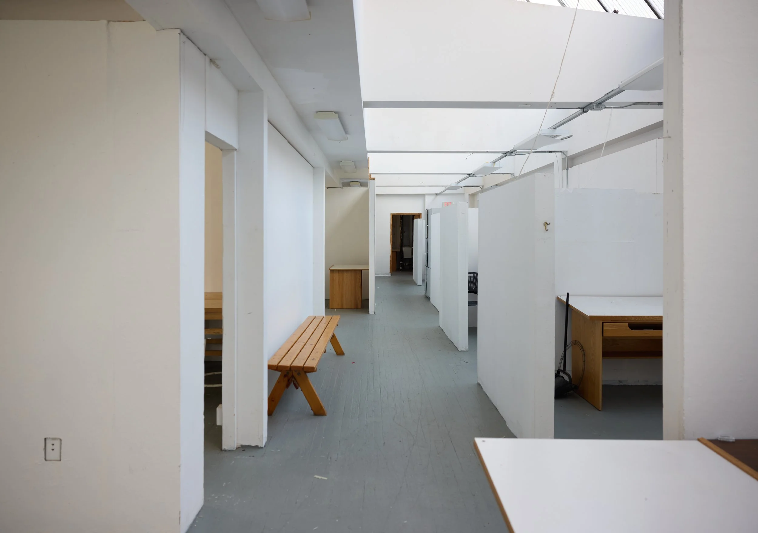 A plain white hallway in one of the buildings at Village of Industry and Art. This photo shows a shared space, divided by cubby walls with a skylight above.