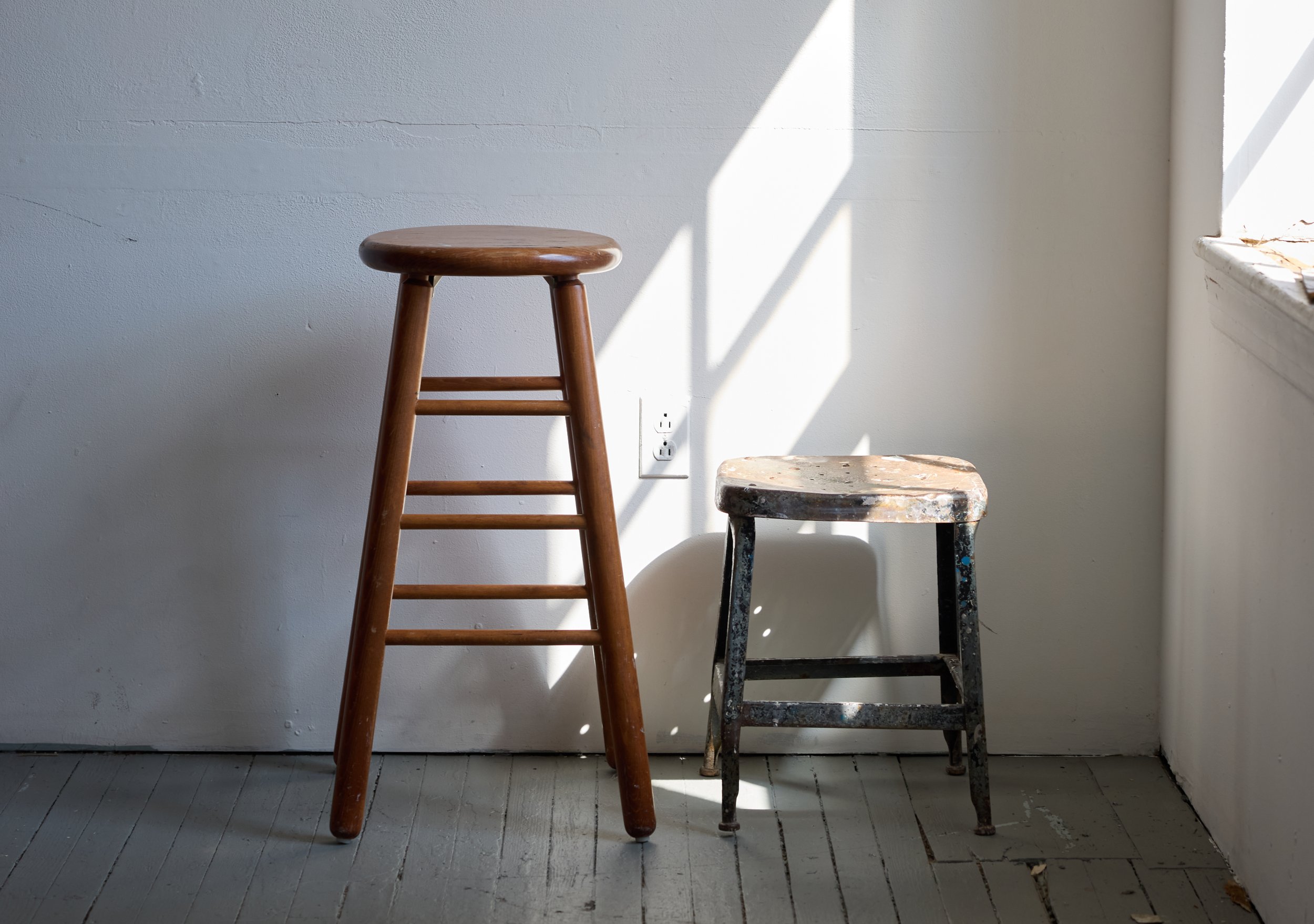 Two stools sitting against a white wall in an empty artist studio space with light shining in from a window on the right.