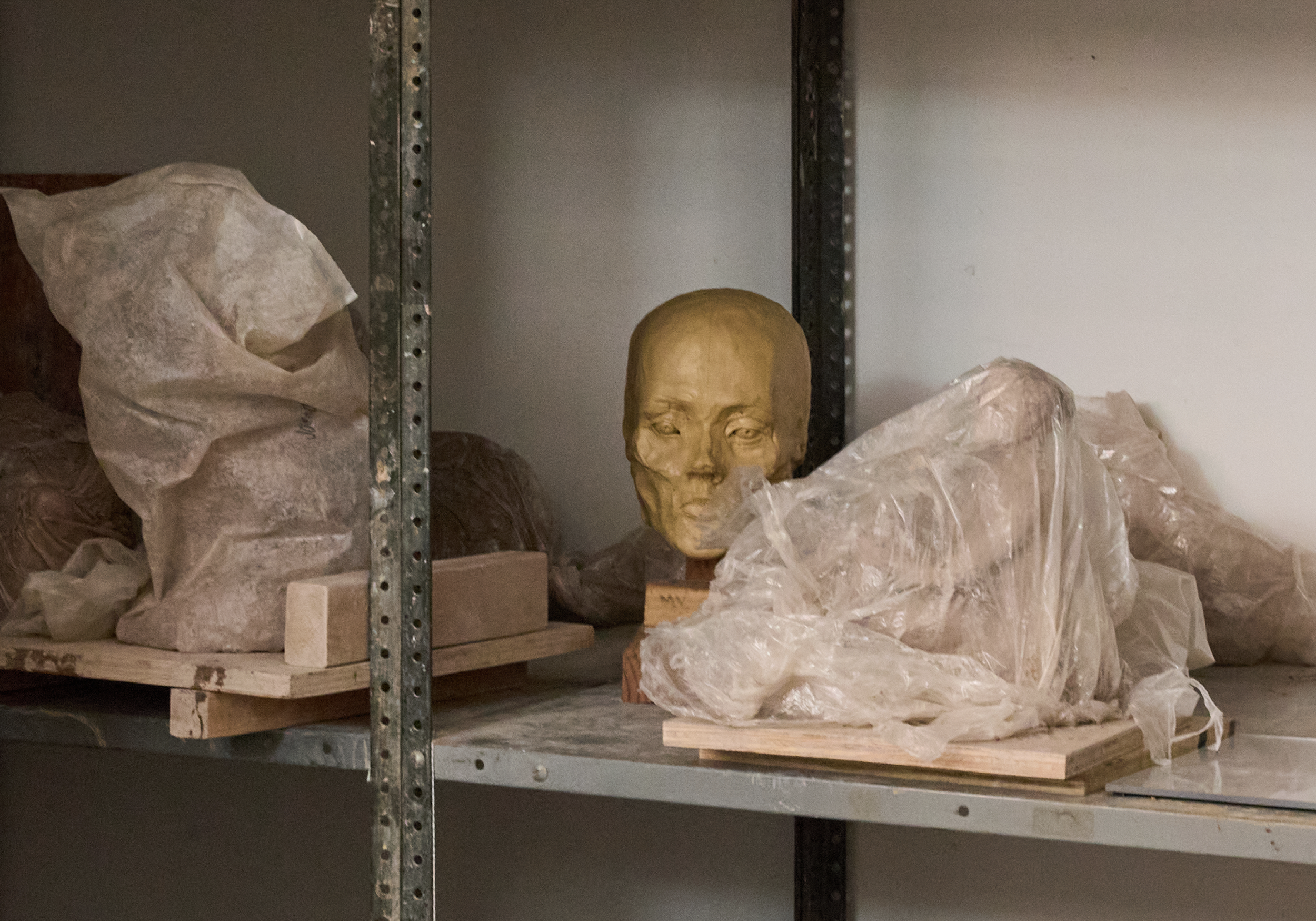 A shelf full of wet wrapped clay projects and a dry molded ceramic human face, representing unfinished work left behind in VIA.