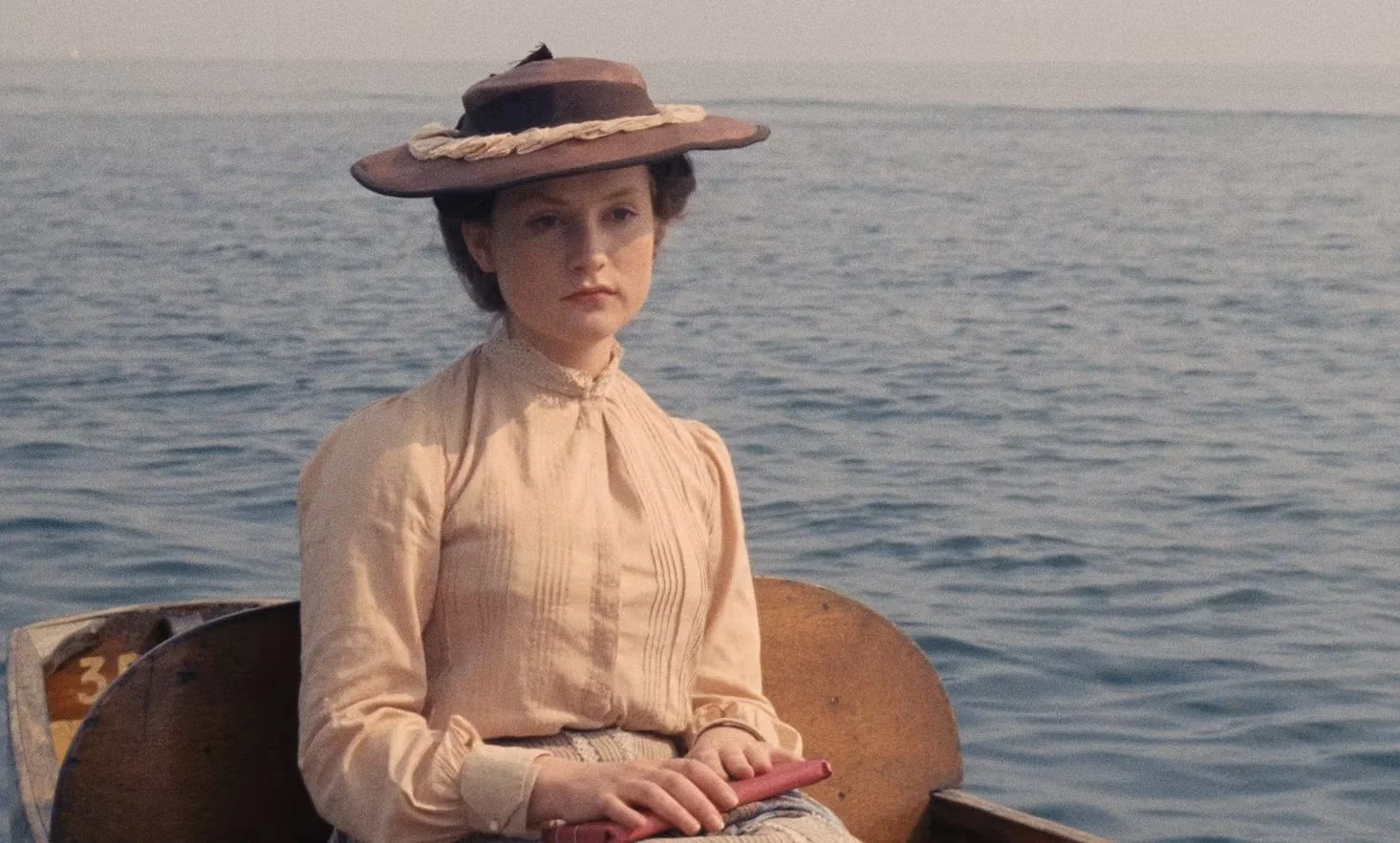 color still image from the film Aloïse depicting a woman in vintage dress seated in a boat against calm rippling water that stretch to the horizon. she is wearing a brown hat and has a serious look on her face.