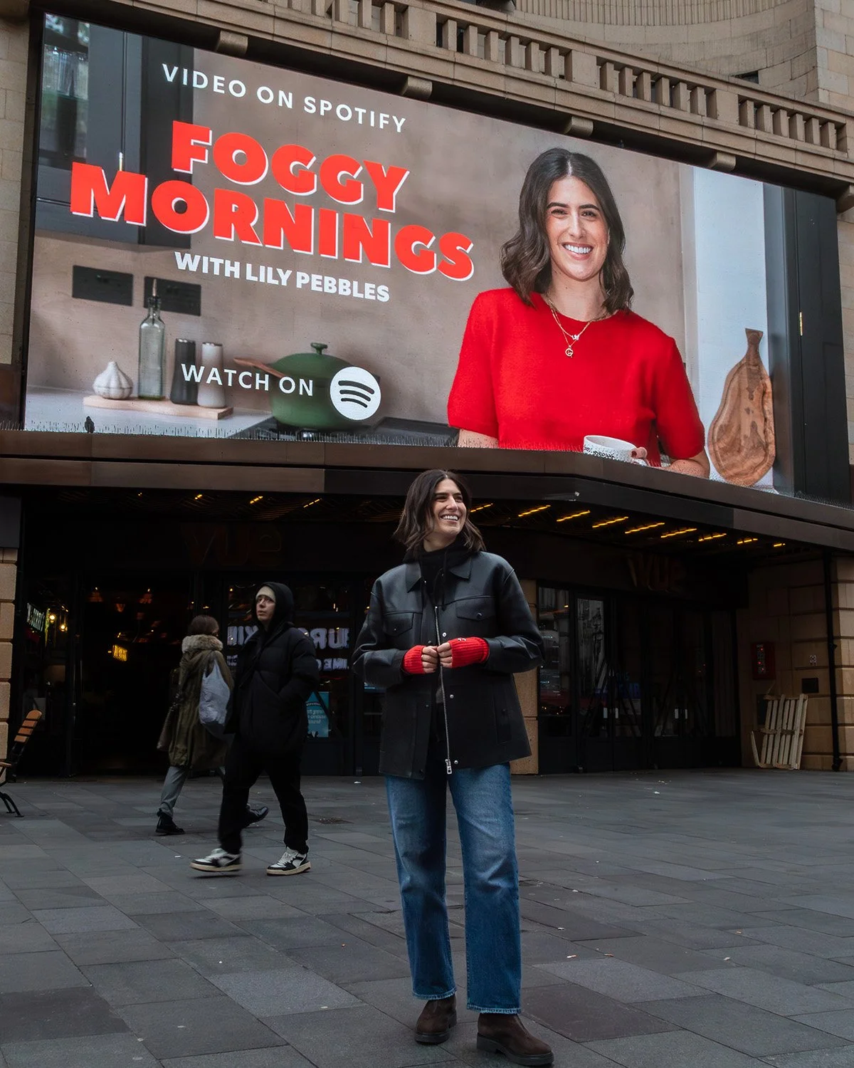 A young woman with shoulder-length brown hair, wearing a black jacket, blue jeans, and dark boots, standing on a city sidewalk, smiling, with a large advertising billboard above her featuring a woman in a red top and the text promoting a video on Spotify called "Foggy Mornings with Lily Pebbles."