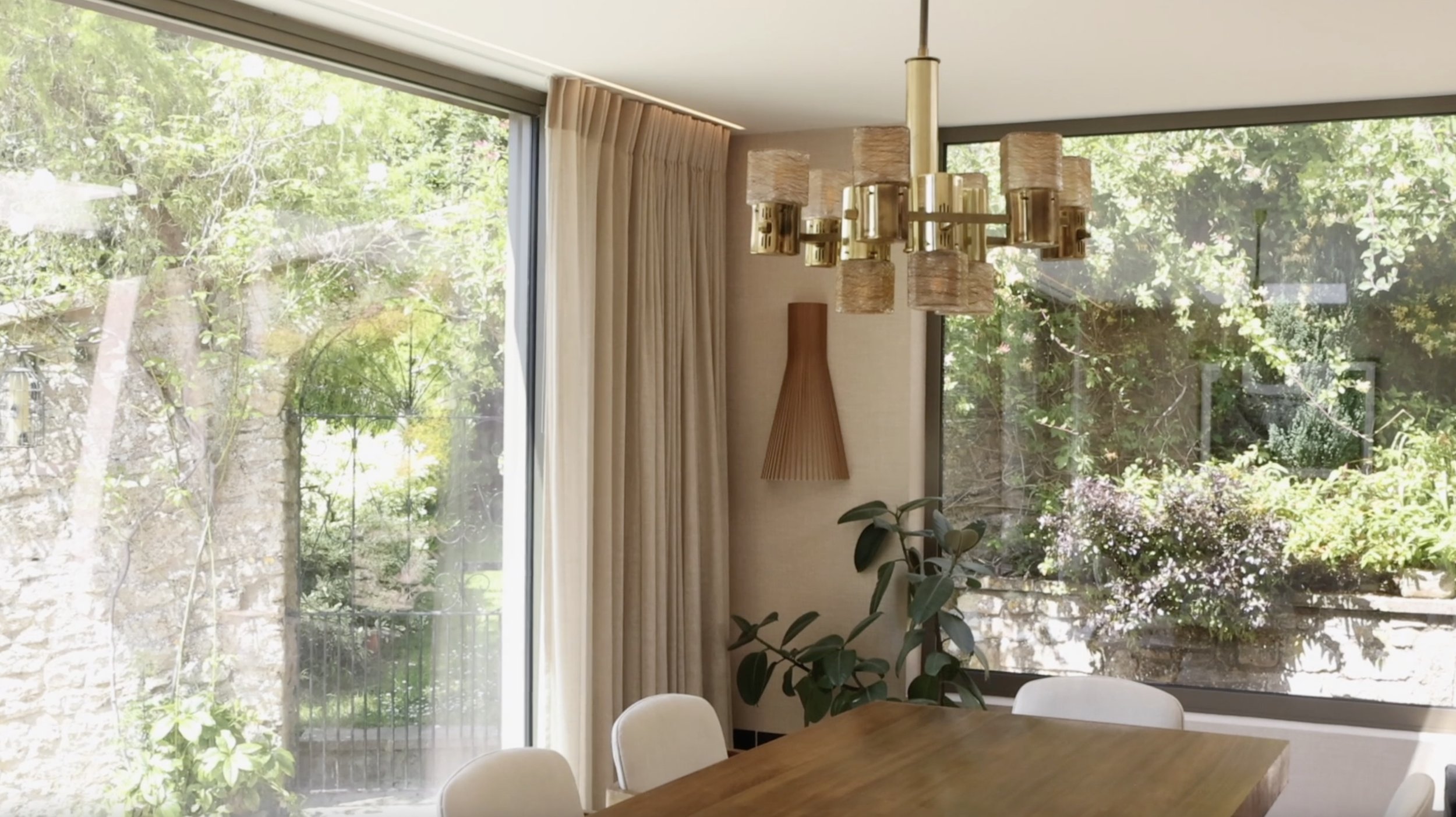 Interior of a dining room with a wooden table, white chairs, a large window, and a view of a garden outside, with beige curtains and a brass chandelier.