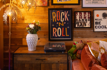 Decorative room corner with a wooden wall, framed wall art, a white textured vase with flowers on a wooden dresser, a record player, plush toys, and pillows on a pink sofa.