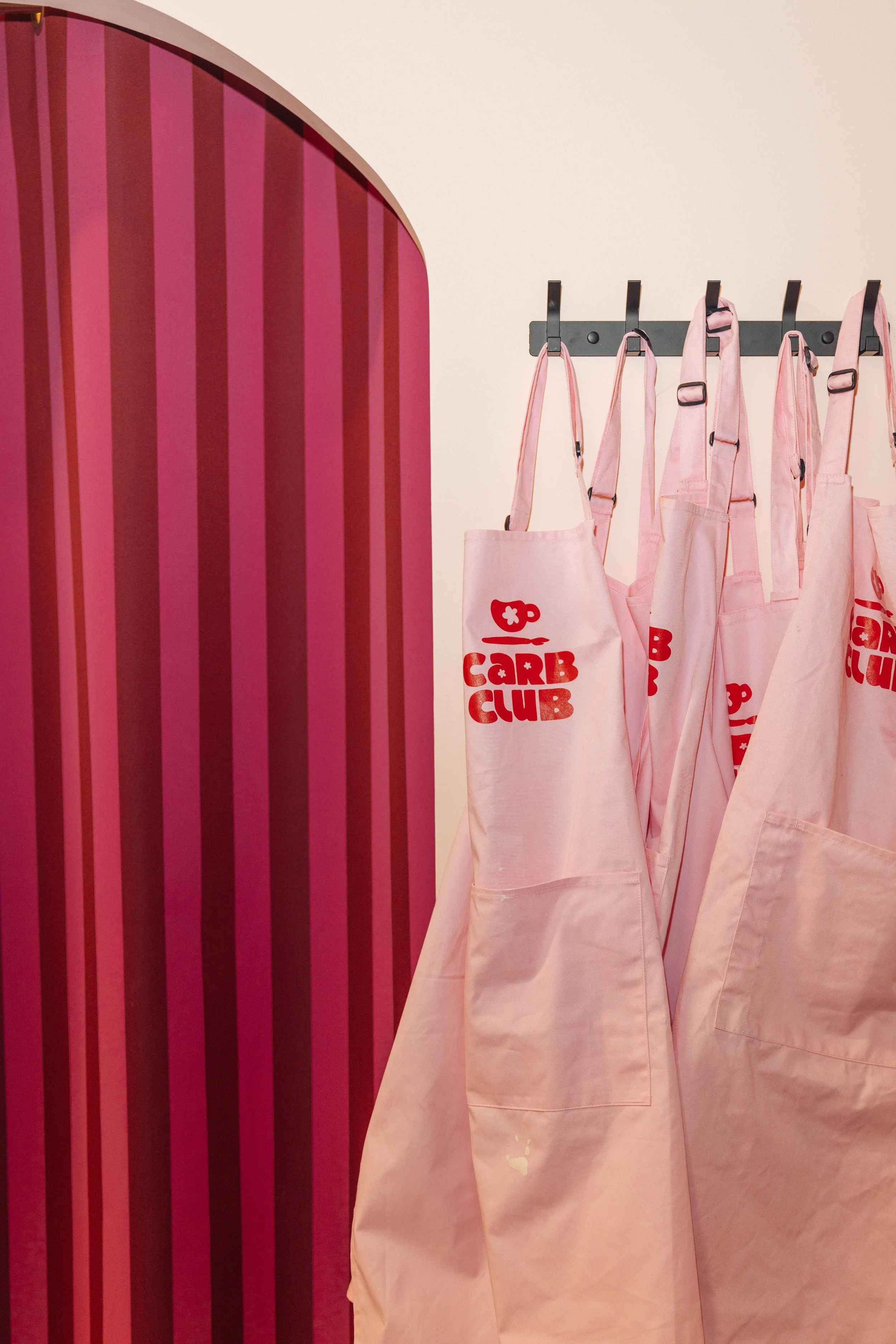 Pink aprons with red text hanging on hooks on a wall, with a red and pink striped background panel.