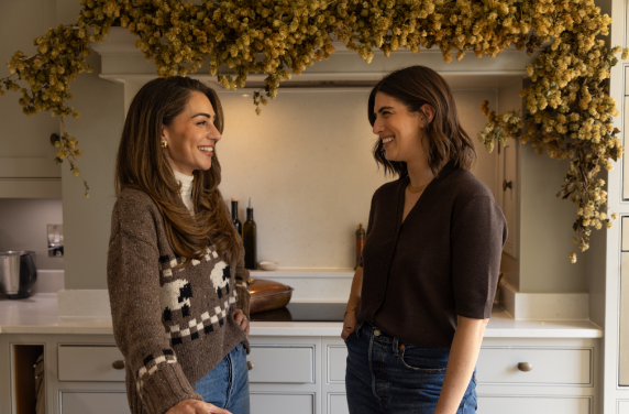 Two women smiling and talking in a kitchen decorated with yellow flowers hanging from the ceiling.