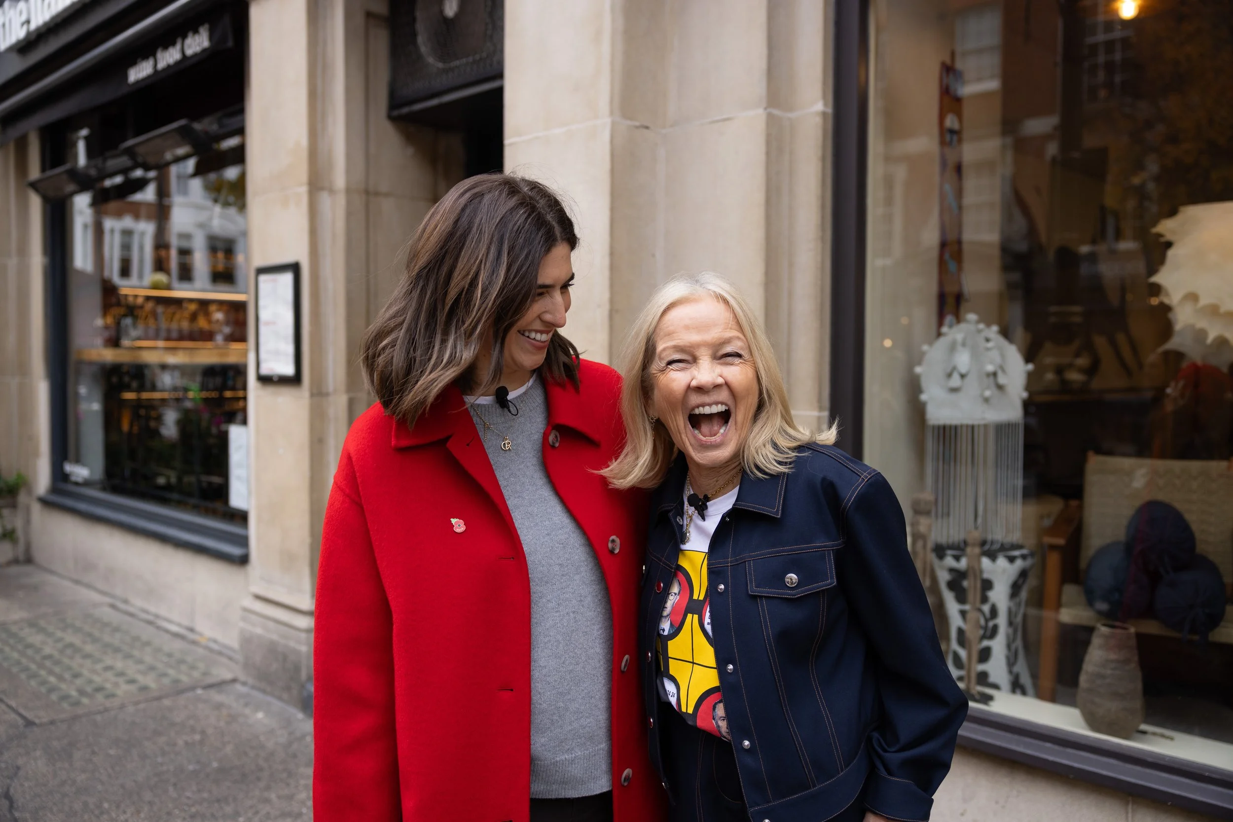 Two women laughing and smiling outdoors in front of a building with large windows, one in a red coat and the other in a denim jacket.