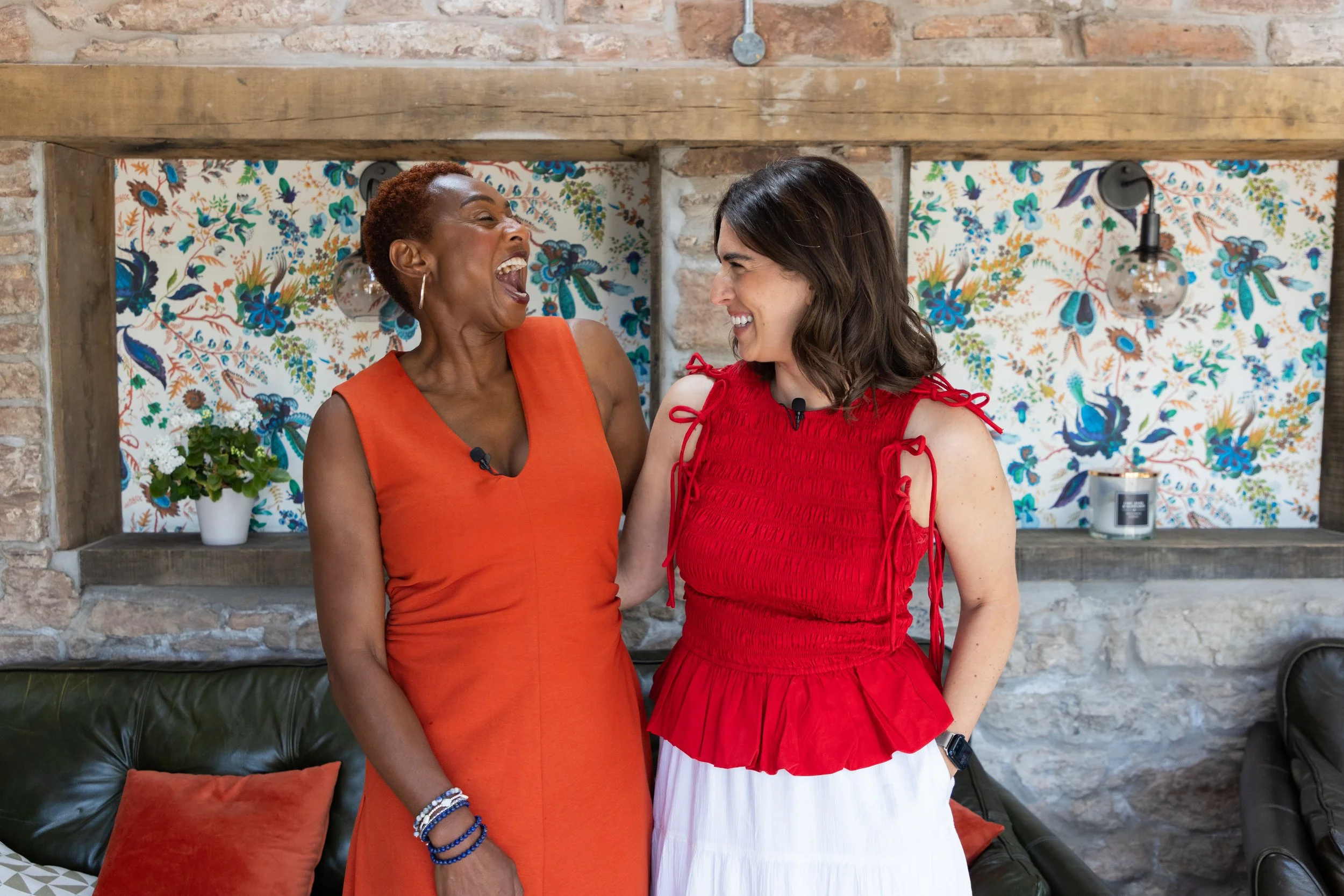 Two women are laughing and smiling at each other indoors. One woman is wearing an orange sleeveless dress, and the other is wearing a red sleeveless top with tied shoulder straps and a white skirt. They are standing in front of a decorative wallpaper with flowers and birds, with shelves holding a potted plant and a candle.