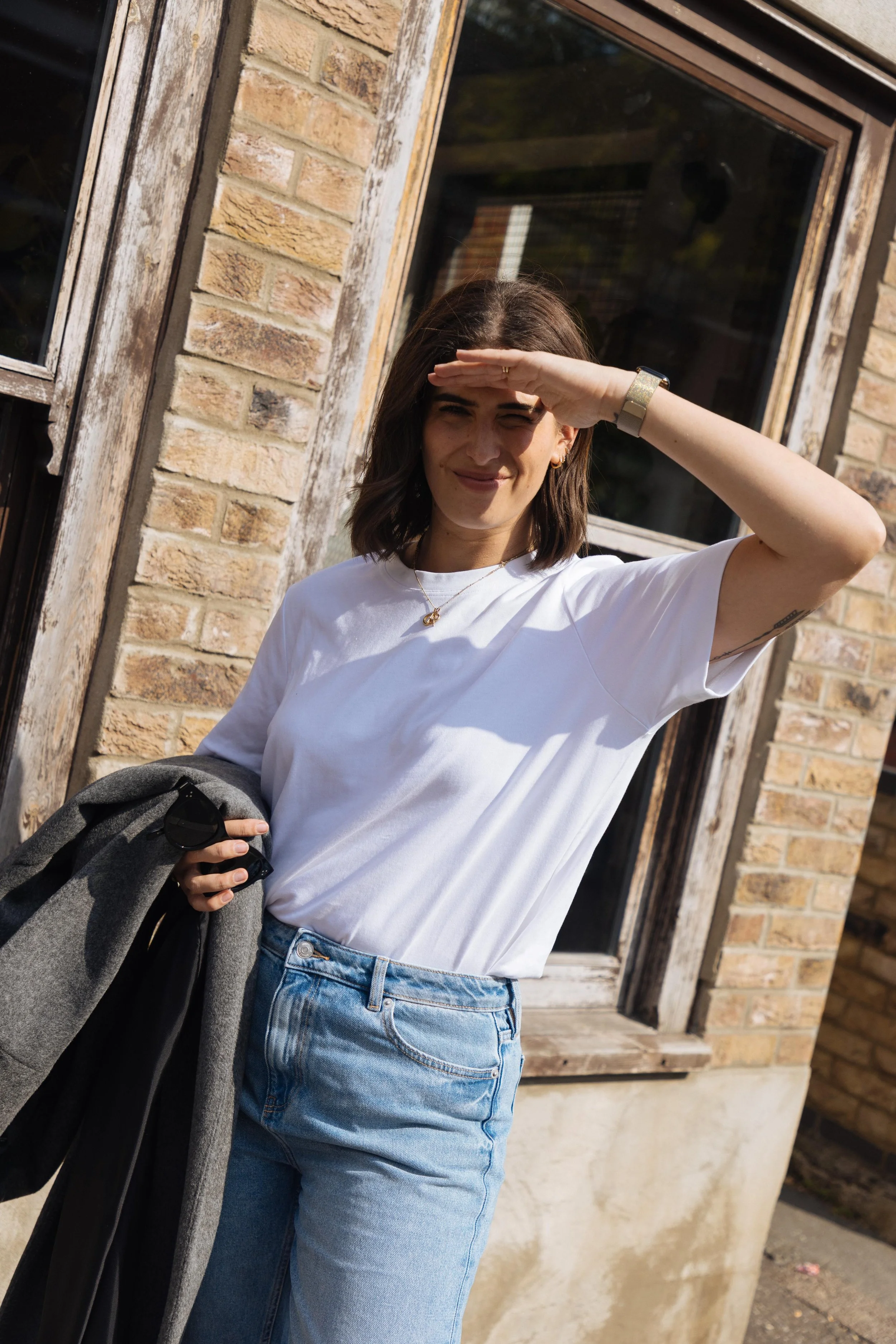 Woman standing outside in front of a brick building, shielding her eyes from the sunlight with her hand, holding a coat and sunglasses in one hand, dressed casually in a white t-shirt and denim jeans.