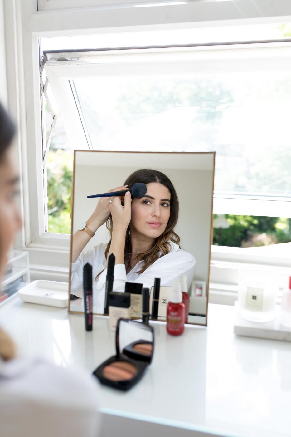 Woman applying makeup in front of a mirror with various cosmetics on the table.