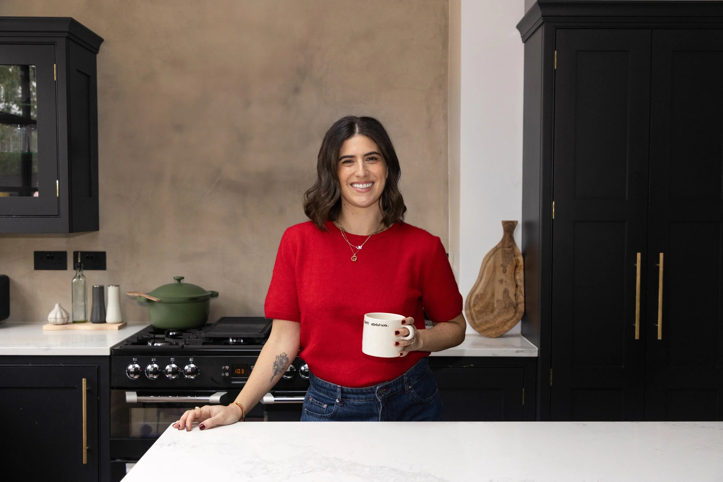 A young woman with shoulder-length dark hair, wearing a red short-sleeve sweater and blue jeans, smiling and holding a white mug in a modern kitchen with black cabinets, a green pot on the stove, and a wooden cutting board in the background.