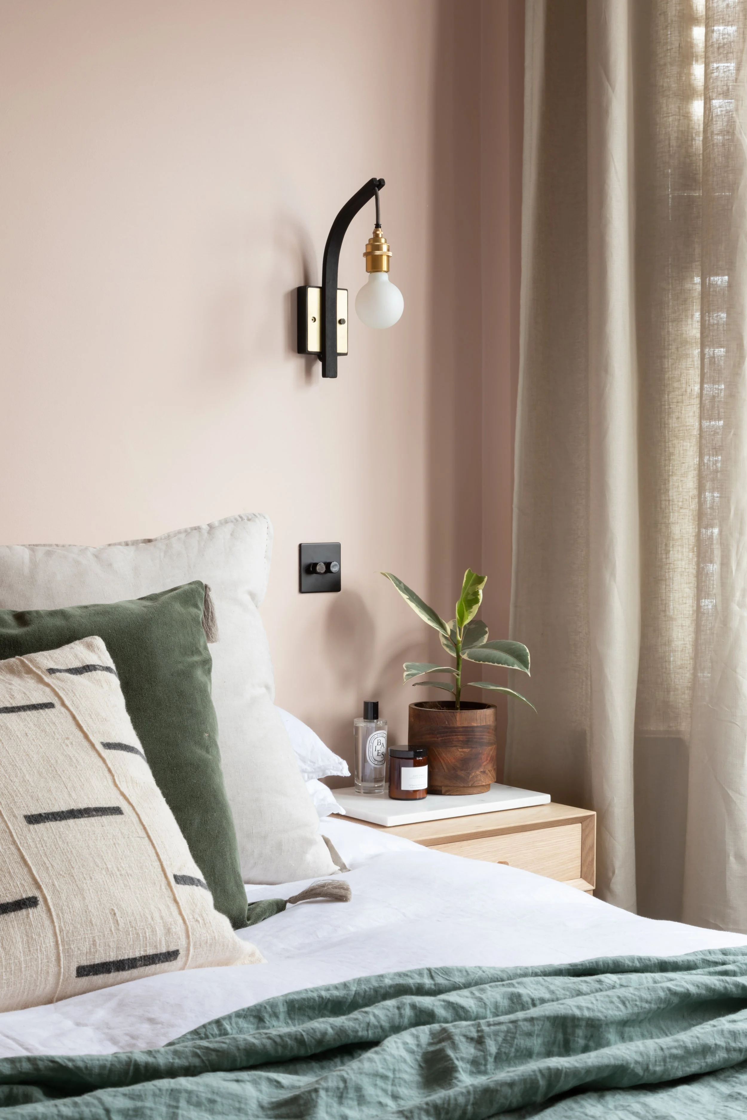 A bedroom with a bed, pillows, a bedside table with a plant, bottles, and candles, a wall sconce, and beige curtains.