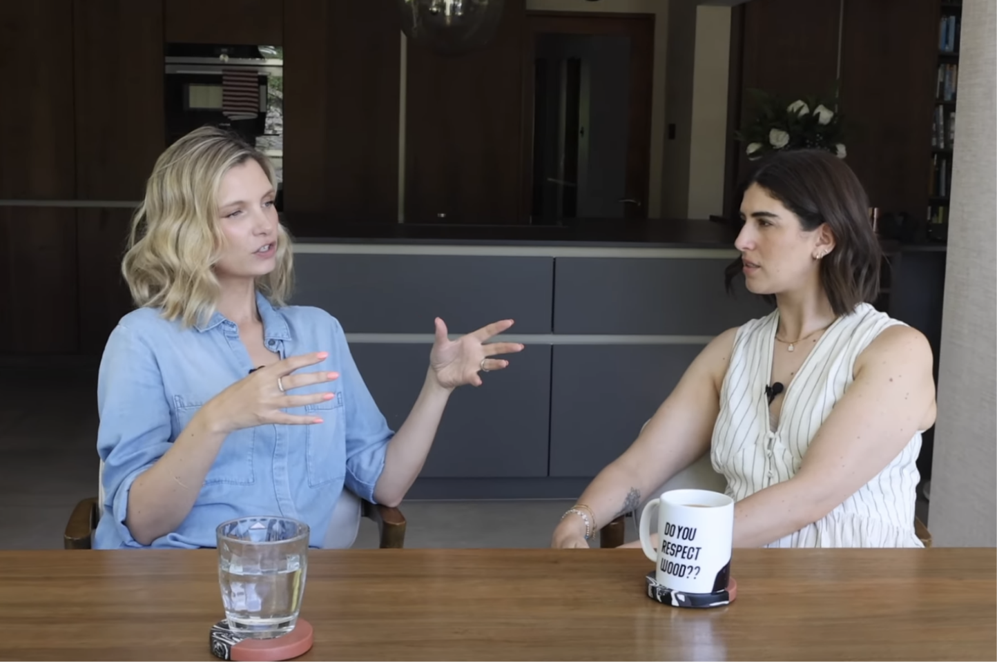 Two women sitting at a wooden table engaged in conversation. The woman on the left has blonde hair and is wearing a light blue shirt, gesturing with her hands. The woman on the right has dark hair, is wearing a sleeveless striped top, and has a mug in front of her that says 'DO YOU RESPECT WOODO??'. There is a glass of water on the table.
