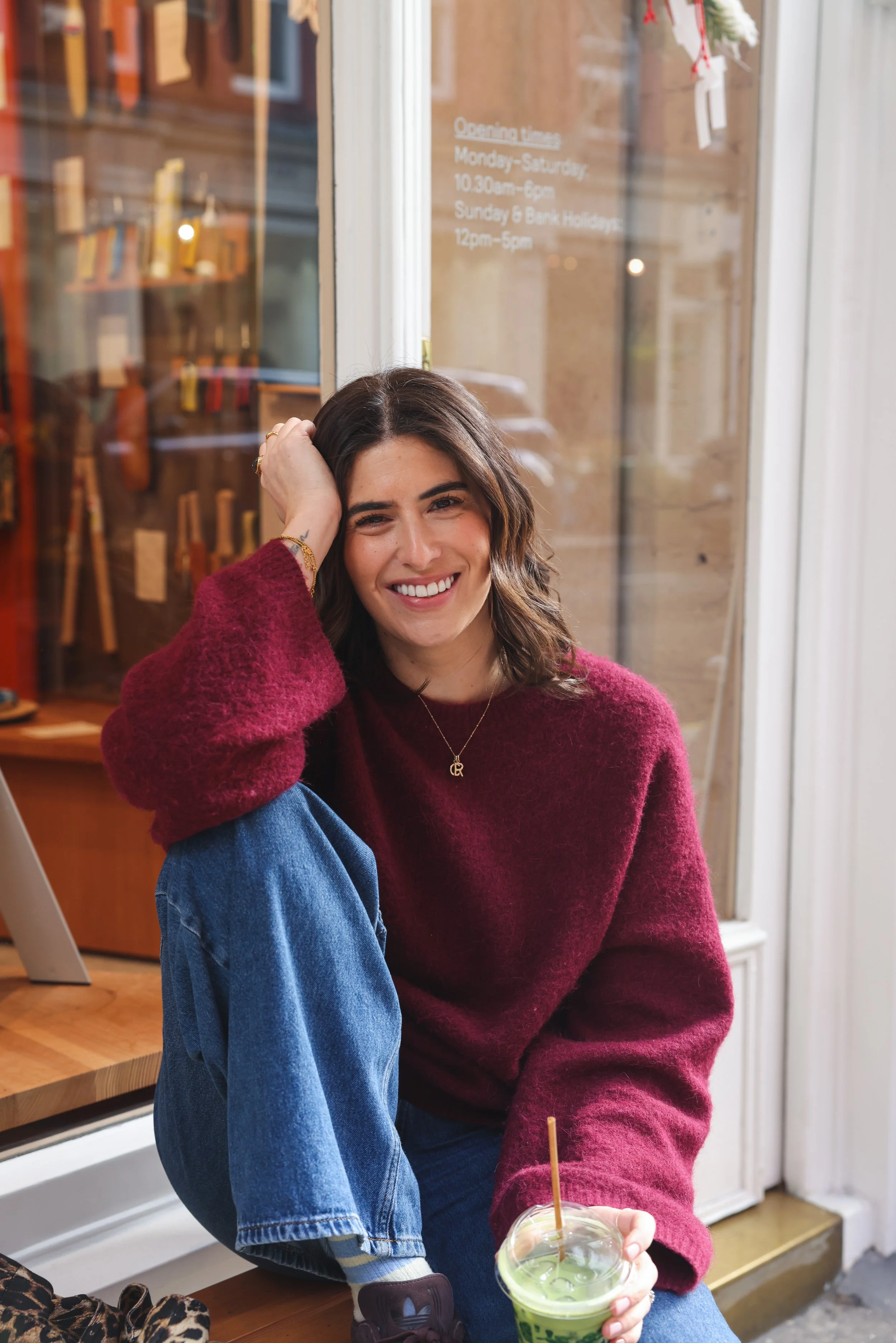 Smiling woman with brown hair, wearing a burgundy sweater and blue jeans, sitting on a windowsill with her left leg up, holding a green iced beverage with a straw, inside a store with retail merchandise visible behind her.