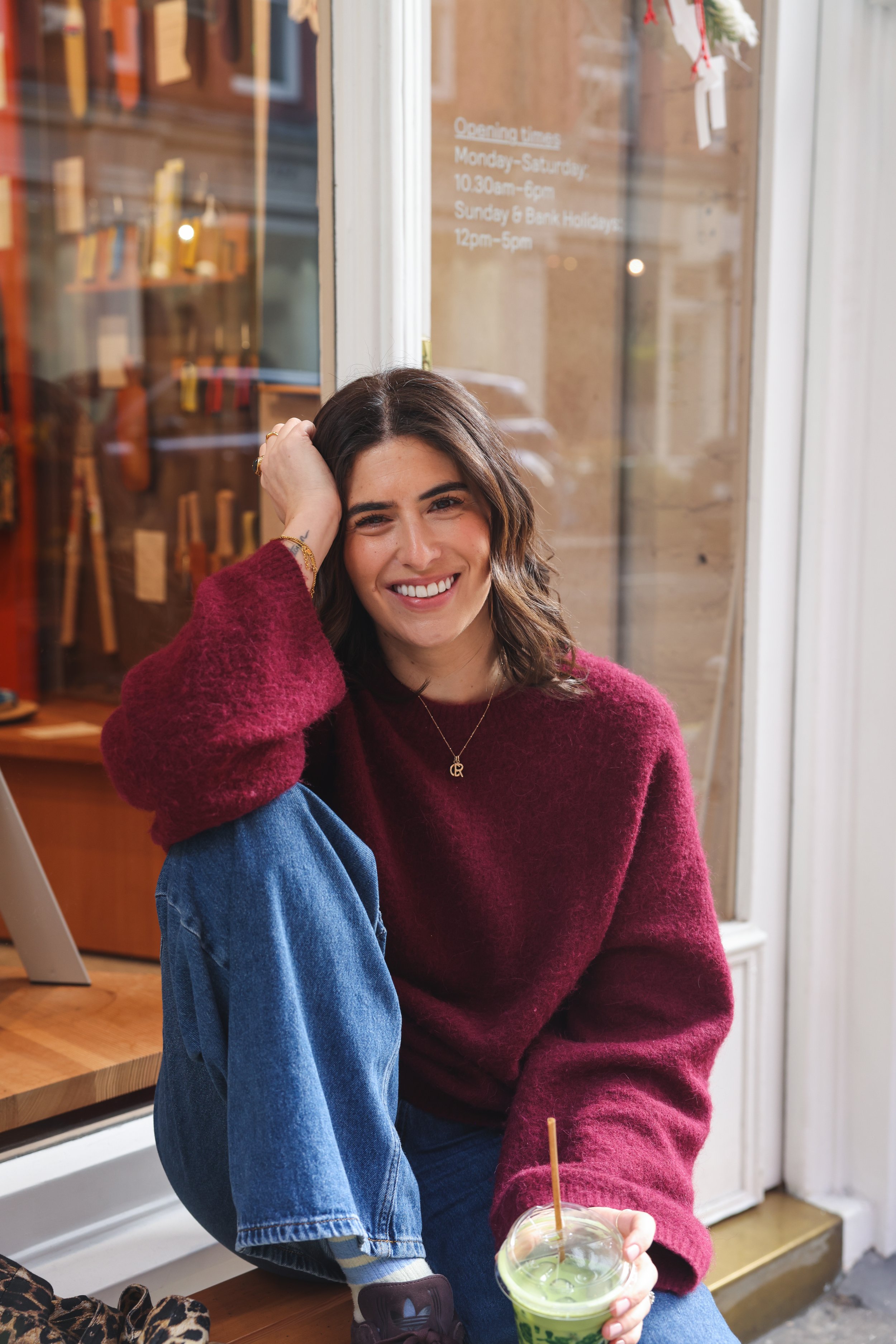 A smiling young woman in a red sweater and blue jeans sitting inside a cafe, holding a green smoothie with a straw, with a display window behind her showing shop hours.