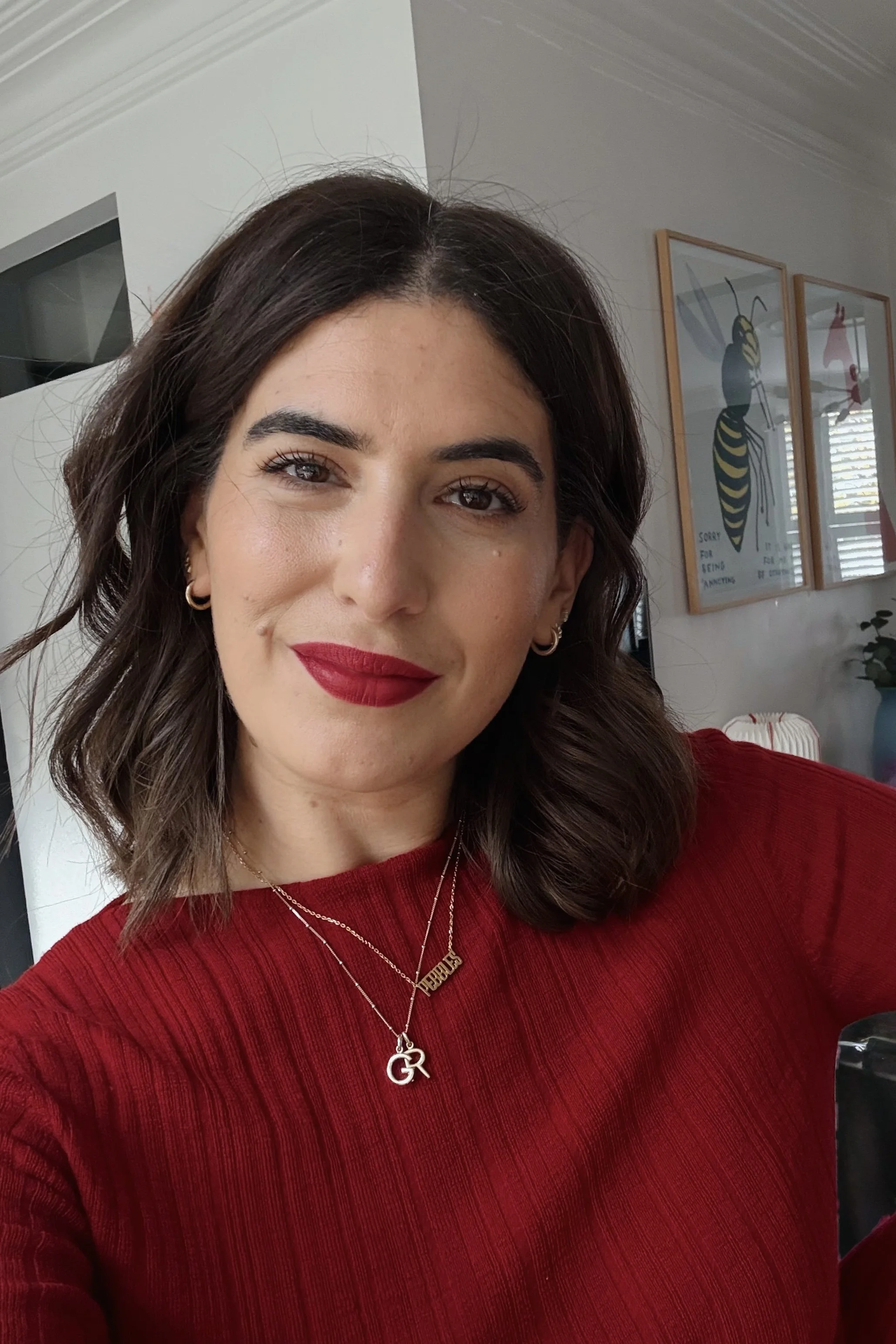 A woman with shoulder-length dark brown hair, wearing a red sweater, red lipstick, and multiple gold necklaces, smiling at the camera in a home interior with framed artwork of bees on the wall.