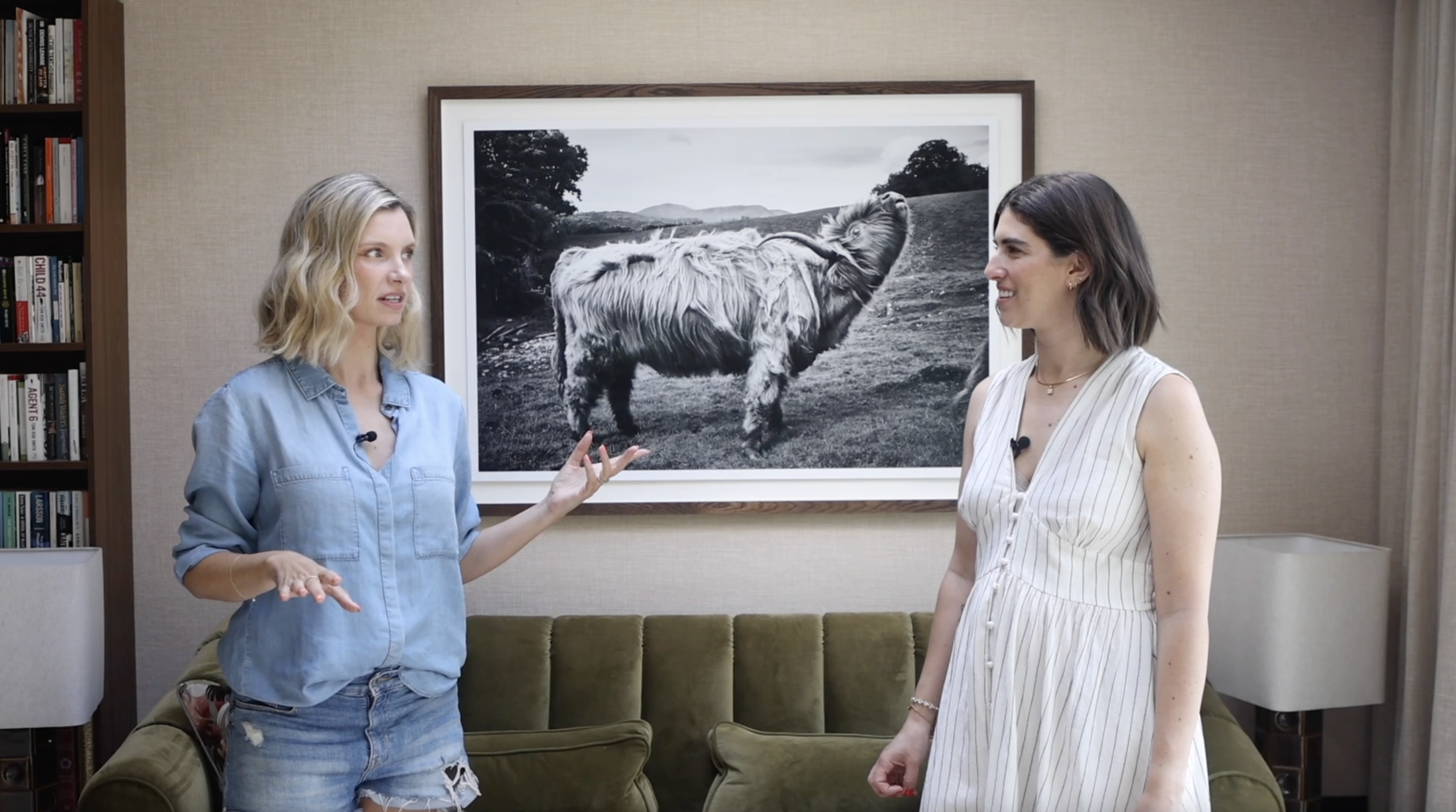 Two women having a conversation in a living room with a large framed black-and-white photograph of a Highland cow hanging on the wall behind them.