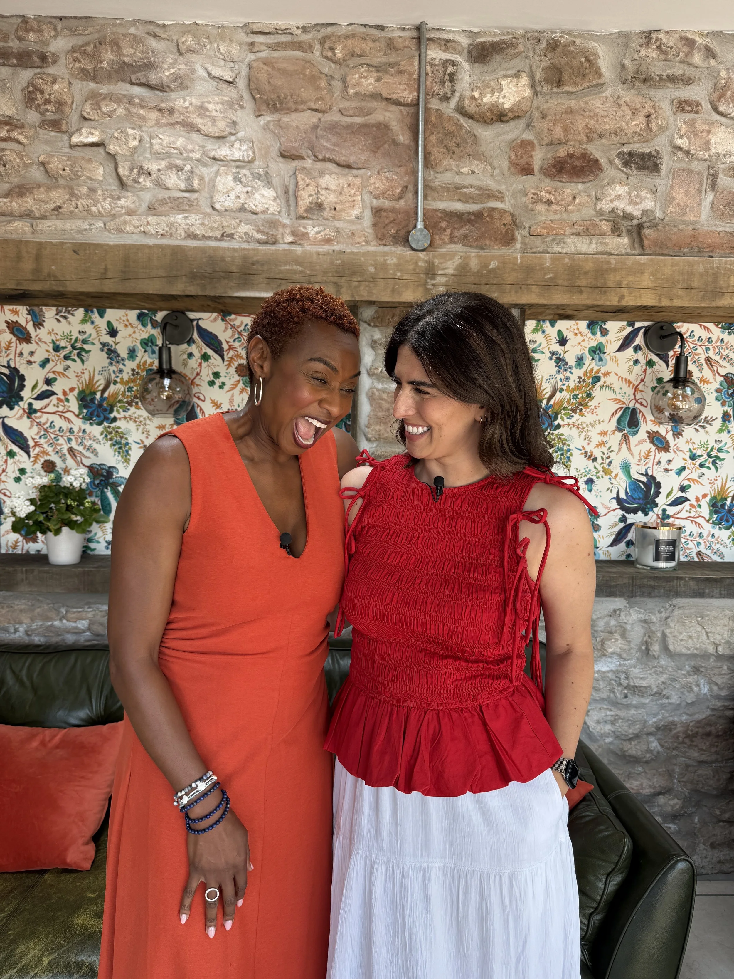 Two women in sleeveless tops, one in orange and the other in red, smiling and looking at each other, standing in a room with a stone wall and floral wallpaper background.