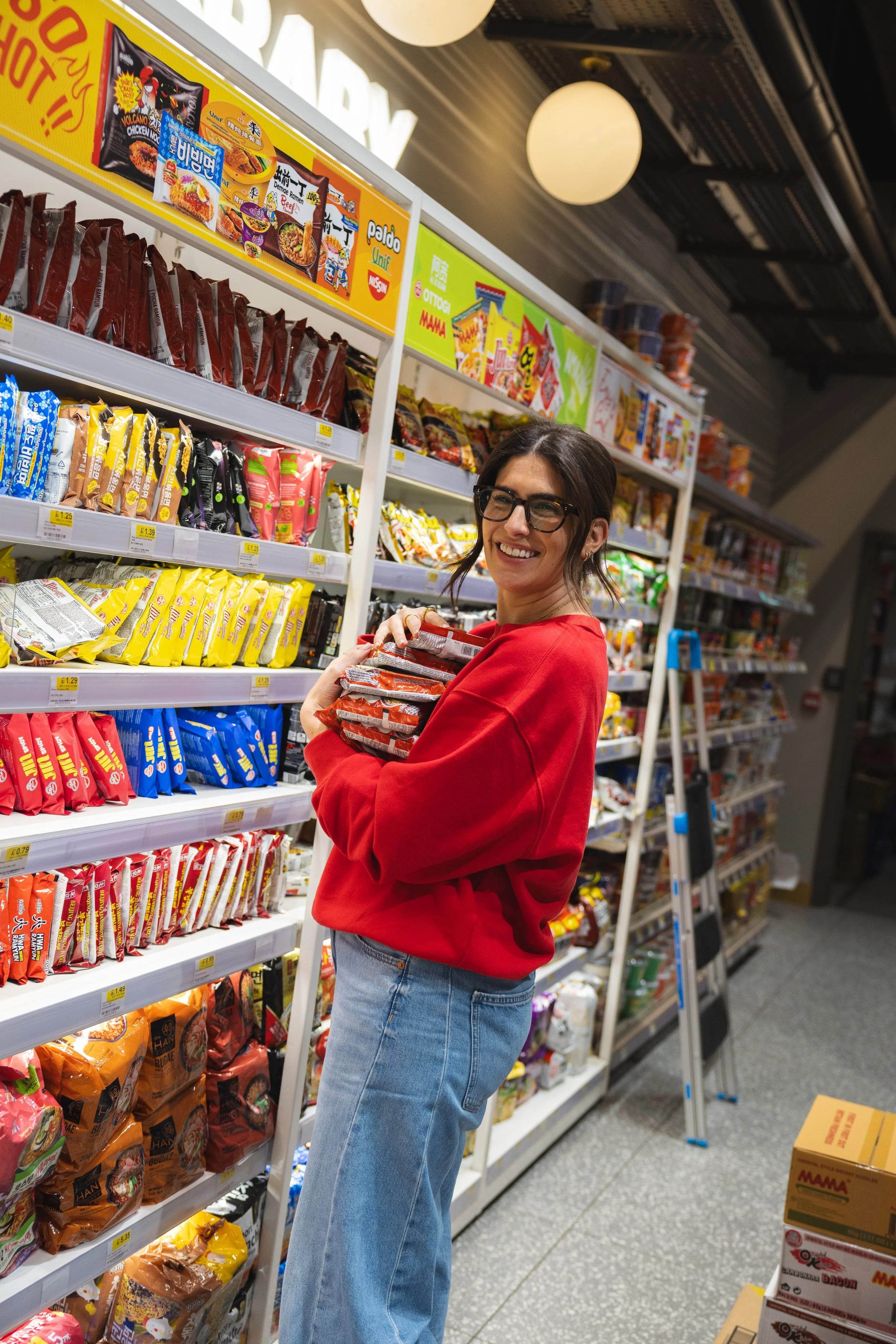 A woman wearing glasses and a red sweatshirt shopping for snacks in a grocery store aisle, holding several packages of instant noodles.